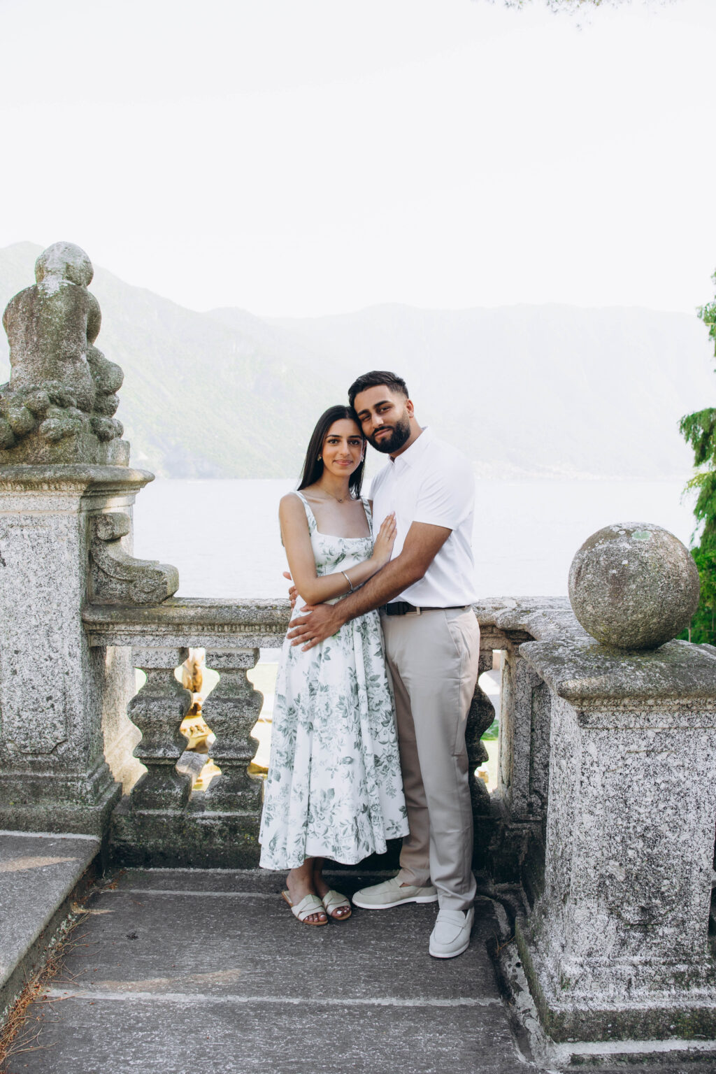 Engaged couple embracing beside a stone cherub statue and a granite sphere on a balustrade above Lake Como in Tremezzo