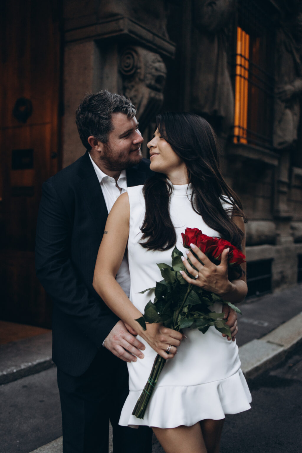 Engaged couple embracing and looking at each other while the woman holds a bouquet of long-stemmed red roses outside the Casa degli Omenoni in Milan