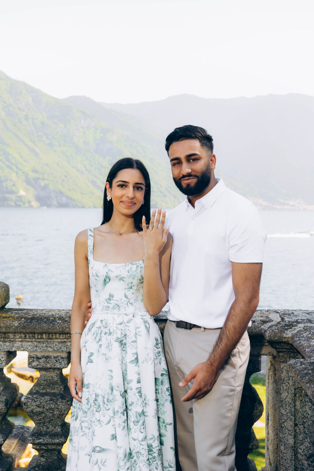 Engaged couple posing at a stone balustrade in Tremezzo as she shows her diamond engagement ring, with Lake Como and mountains behind