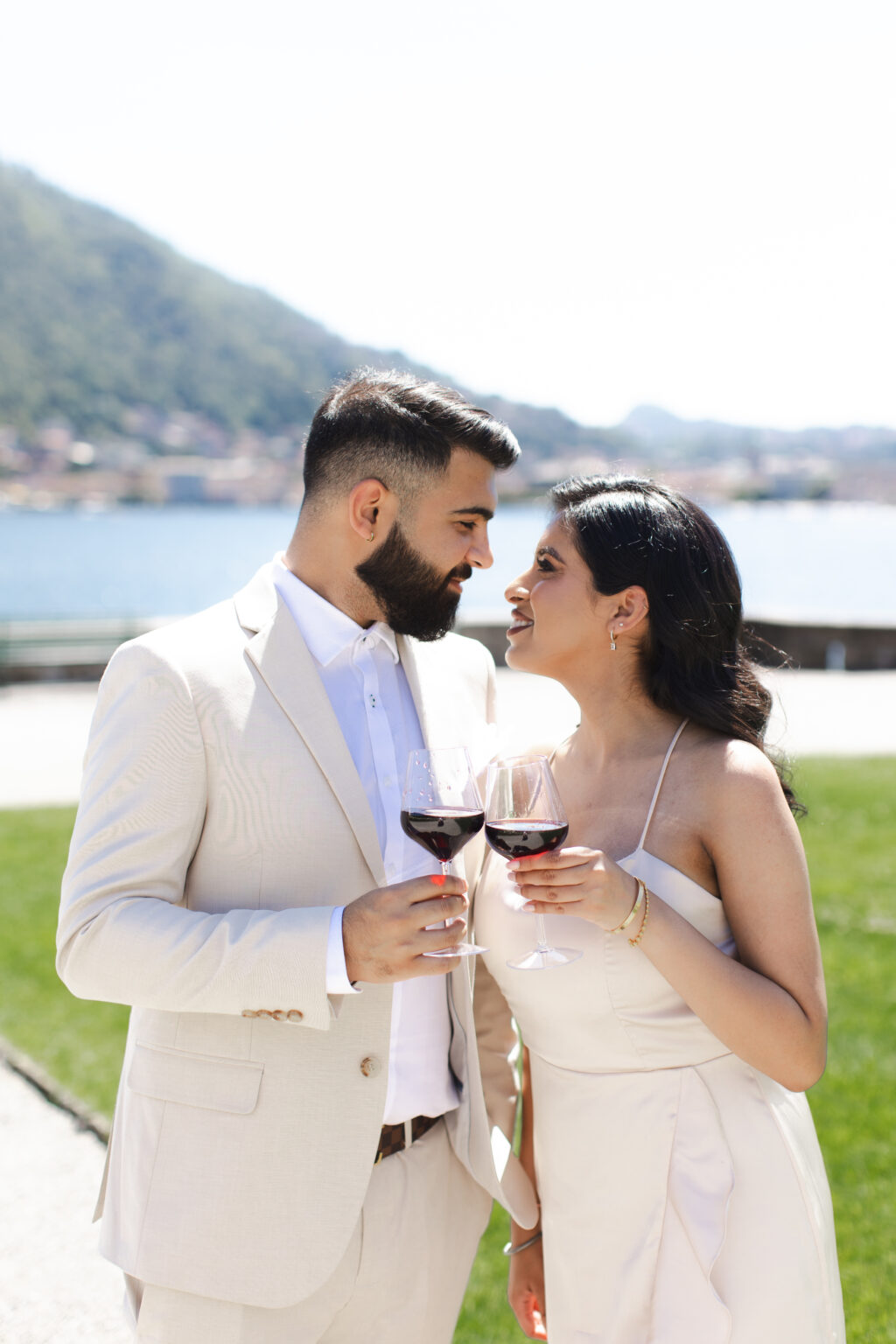 A newly engaged couple standing face to face with red wine glasses, smiling at each other on a lakeside lawn on Lake Como.
