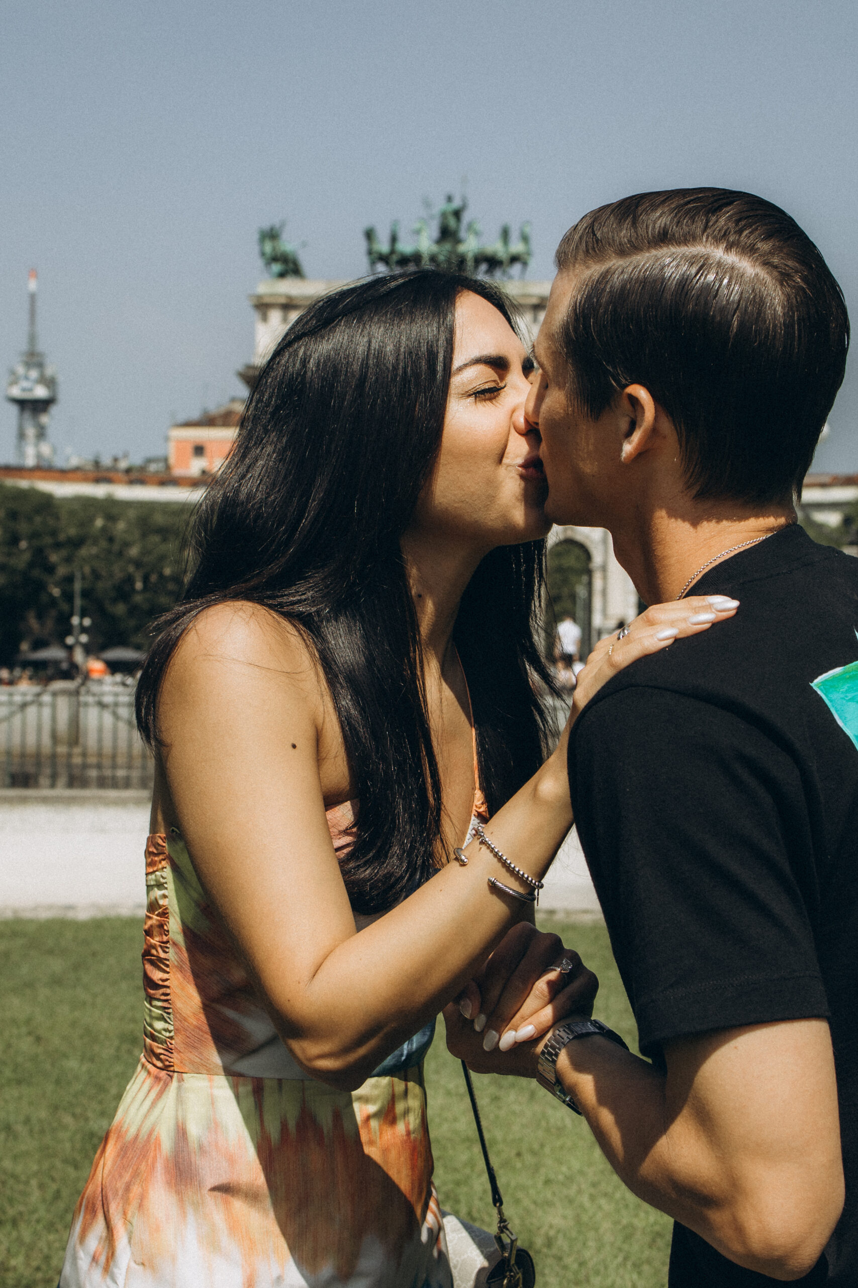 An engaged couple shares a kiss on a sunny day with the Arco della Pace triumphal arch behind them in Milan