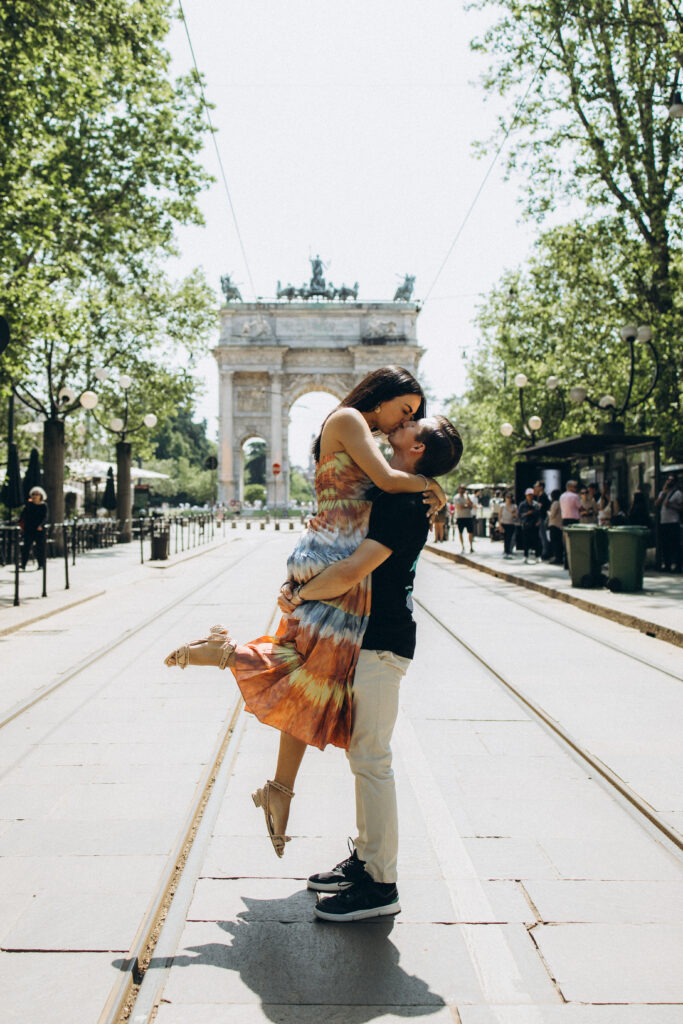 A man lifts his partner into a kiss on a tree-lined avenue with tram tracks leading to the Arco della Pace in Milan