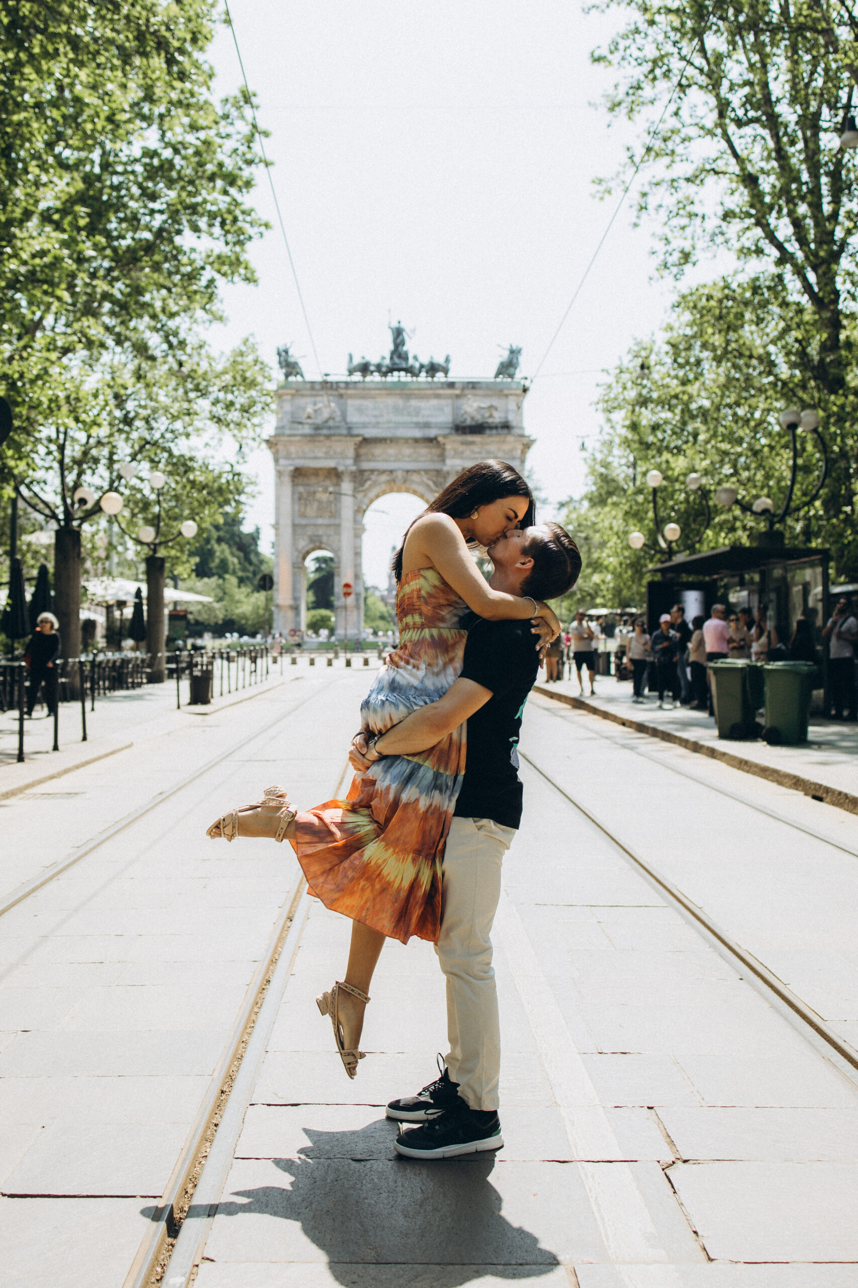 A man lifts his partner into a kiss on a tree-lined avenue with tram tracks leading to the Arco della Pace in Milan