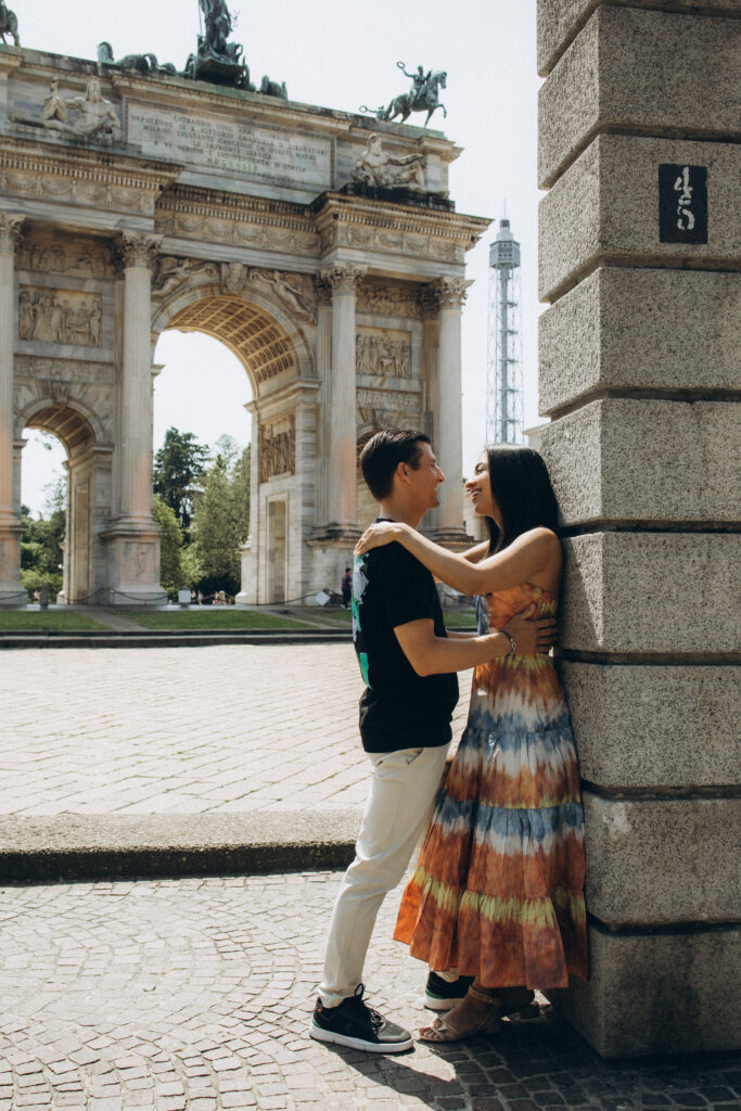 Engagement Photoshoot at Arco della Pace, Parco Sempione, Milan An engaged couple embraces beside a stone pillar with the Arco della Pace and Torre Branca in the background in Parco Sempione, Milan
