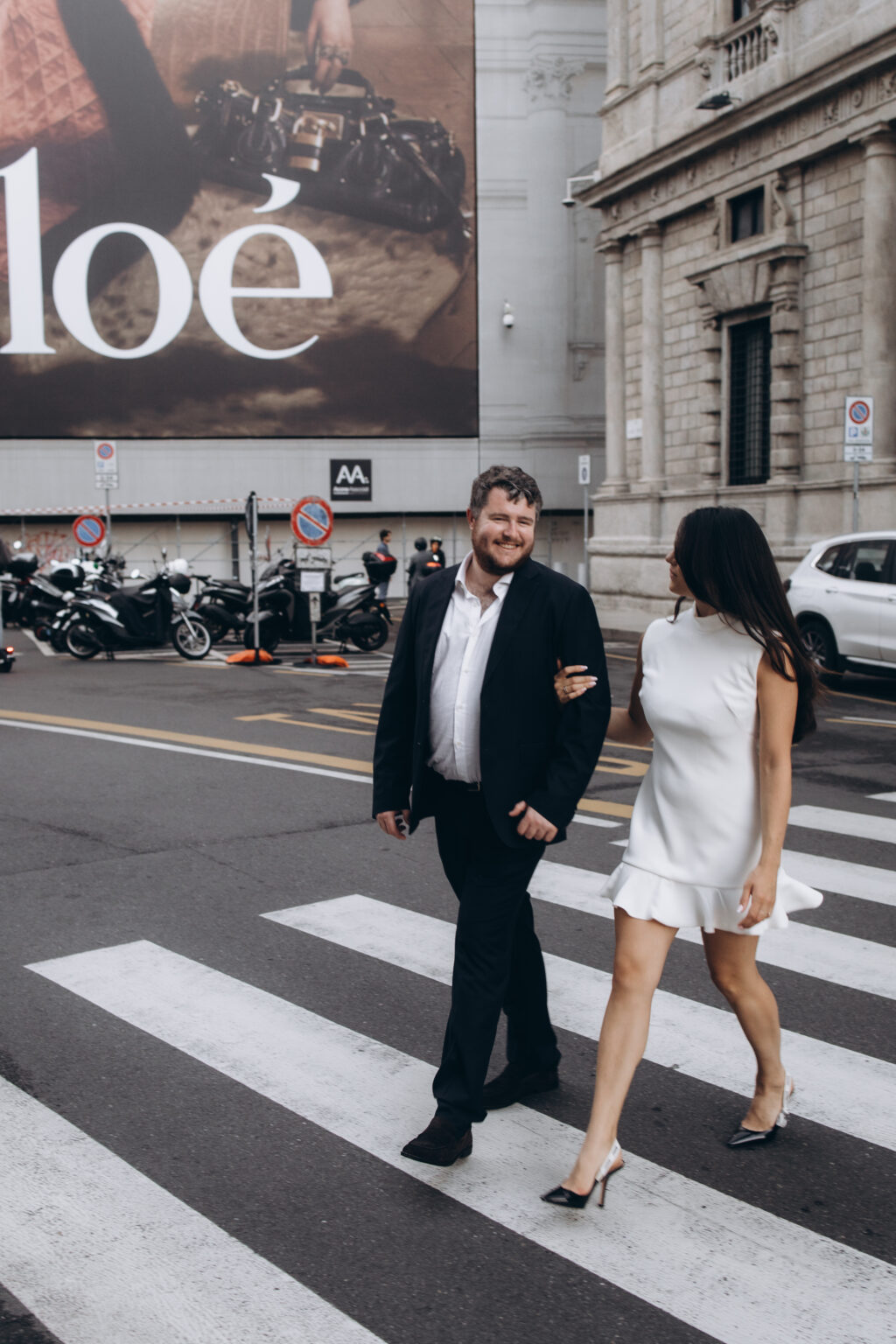 Smiling groom-to-be and his fiancee crossing a Milan street on a pedestrian crossing, with a large fashion billboard on the building behind them