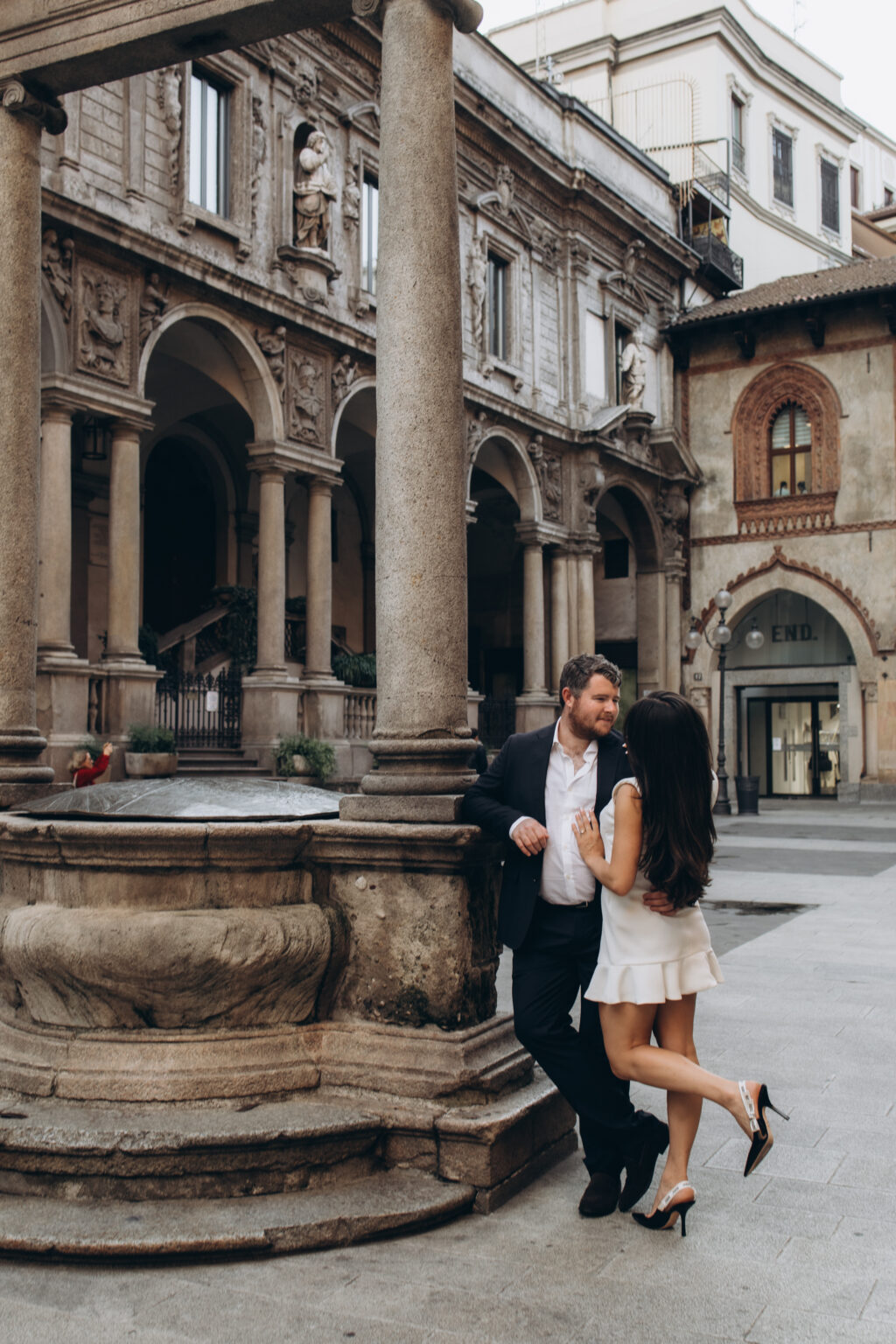 Engaged couple standing close beside the old stone well of Piazza dei Mercanti, with the statue-lined loggia of the Palazzo dei Giureconsulti behind them, in Milan