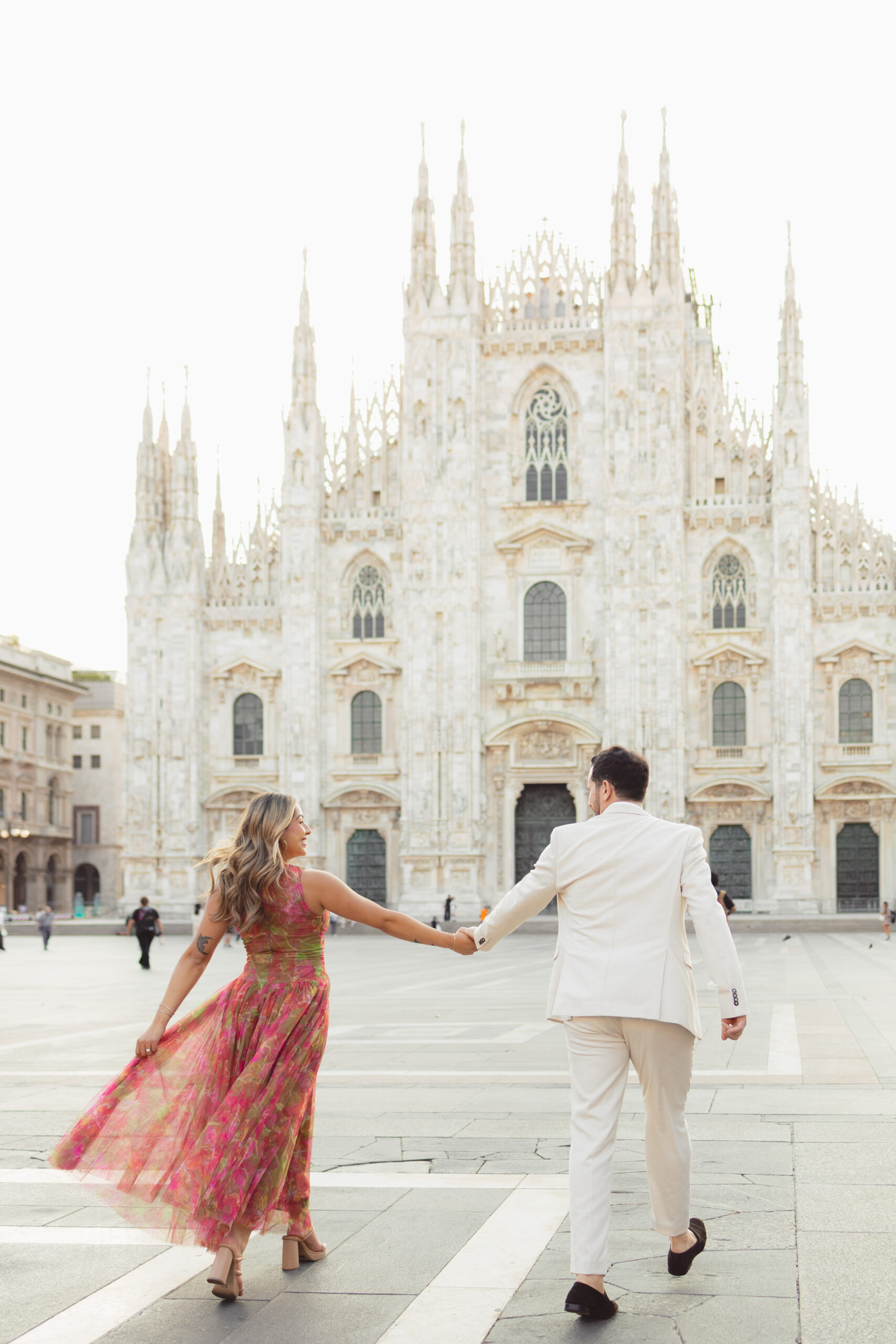 A newly engaged couple walks hand in hand toward the Duomo cathedral across an empty Piazza del Duomo in Milan at sunrise
