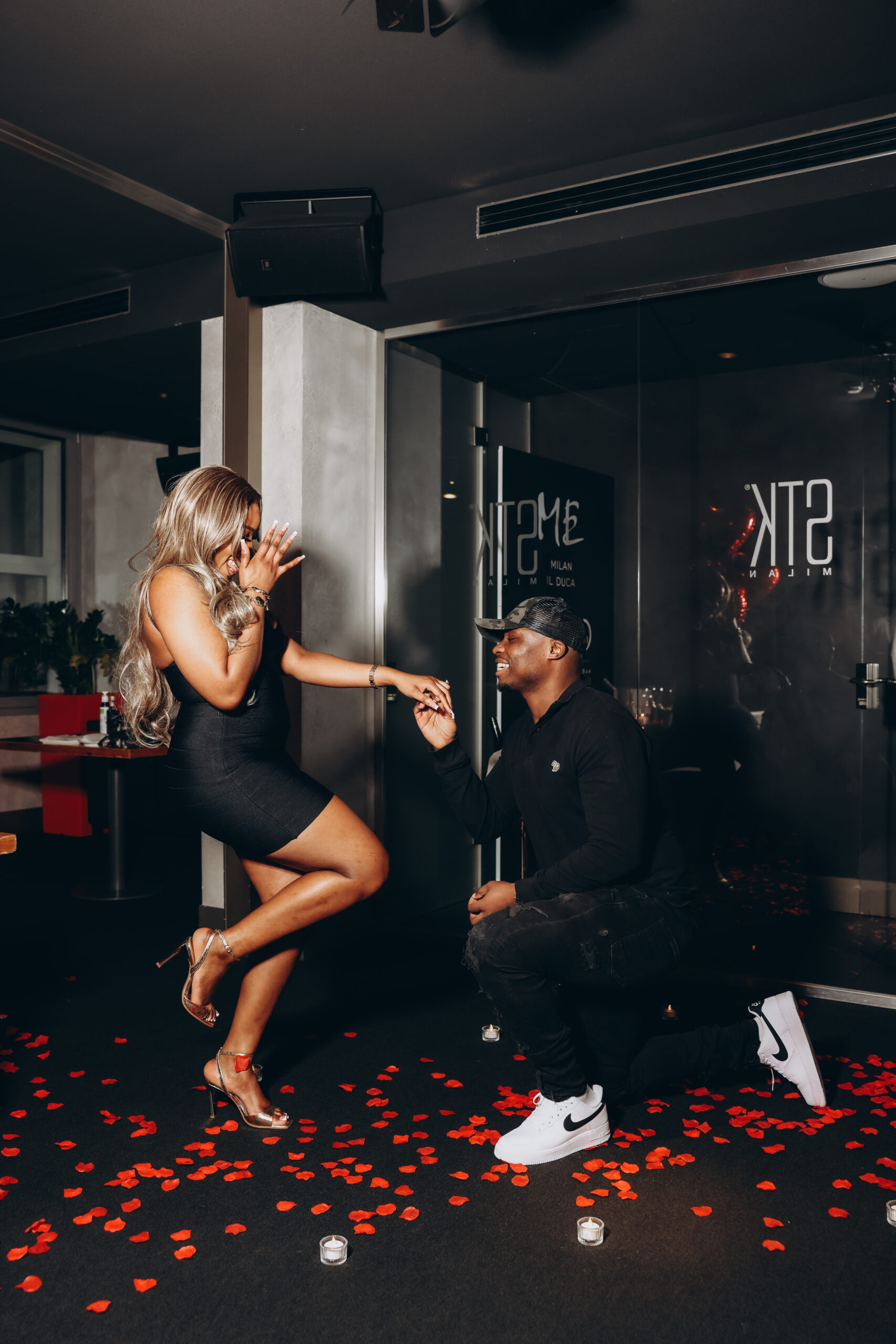 An overjoyed woman covers her face as her partner kneels and holds her hand during a proposal in Milan