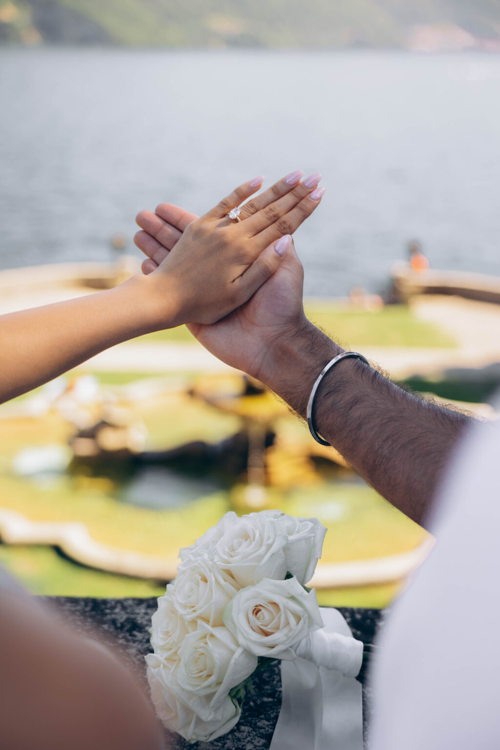 Close-up of the couple's hands with an oval diamond engagement ring above a white rose bouquet, with the fountain of Parco Teresio Olivelli blurred below