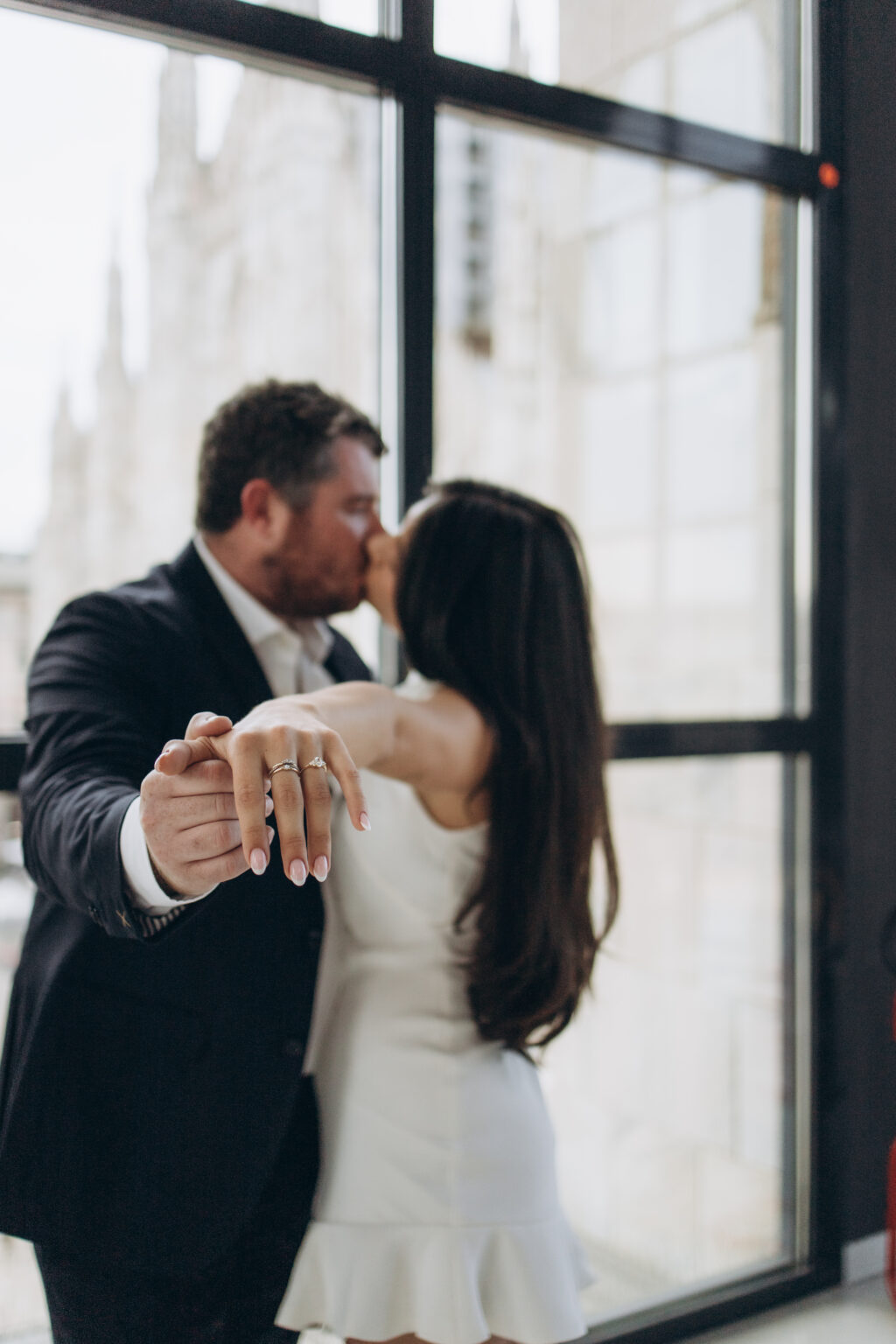 Newly engaged couple kissing as the bride-to-be shows her engagement ring, with the Duomo seen through the Museo del Novecento window in Milan