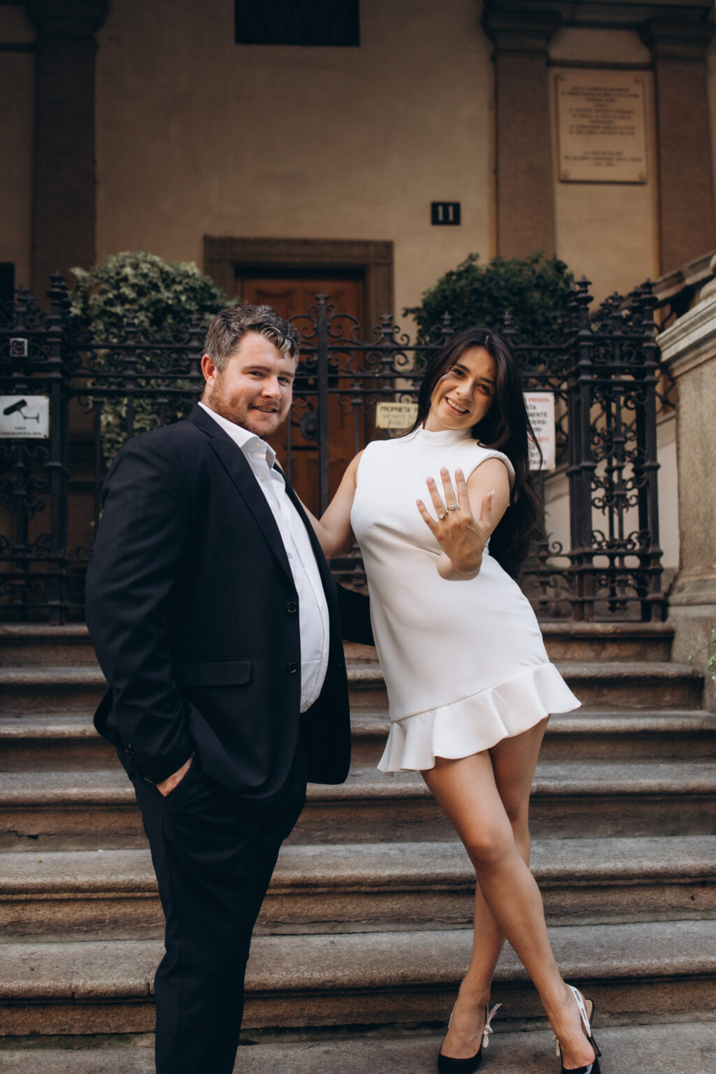 Smiling woman showing her engagement ring to the camera while standing beside her proud fiance in a Milan palazzo courtyard