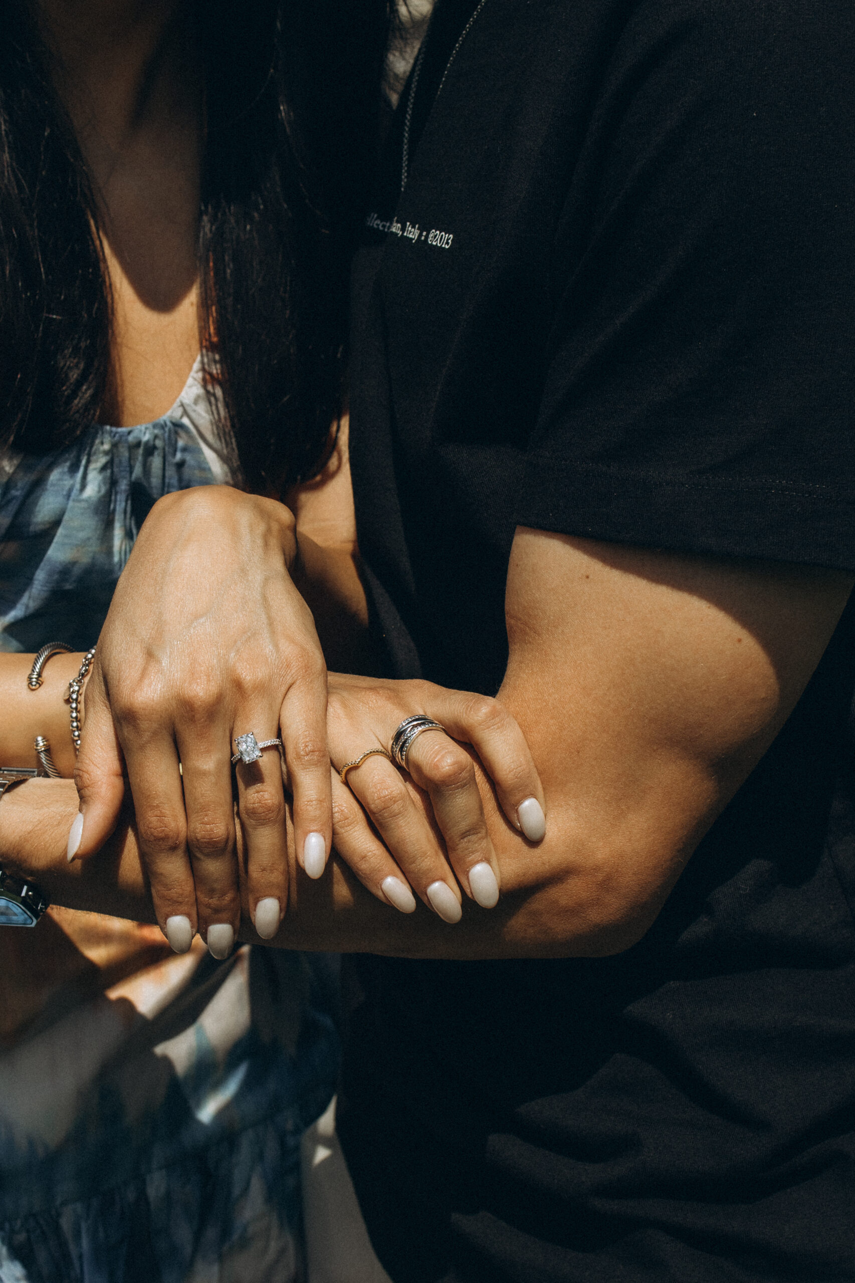 Close-up of a couple's hands resting together in sunlight, showing the woman's solitaire engagement ring and the man's rings in Milan