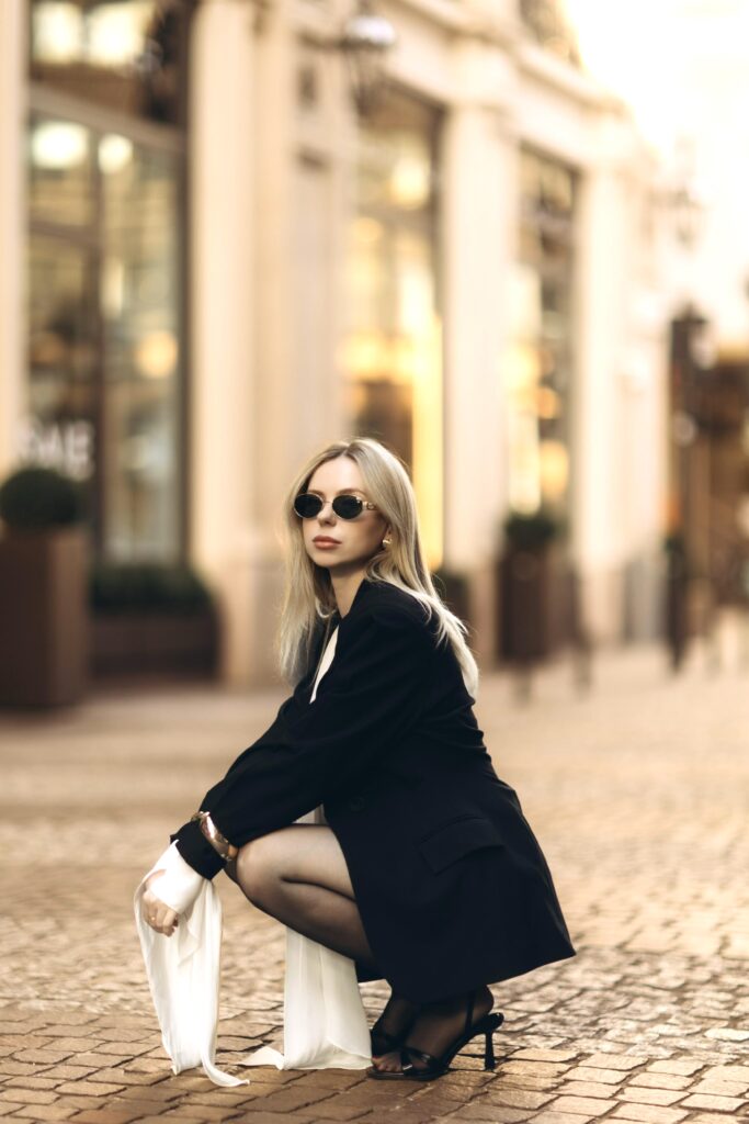 Woman in an oversized black blazer, white blouse, and oval sunglasses crouching on a cobbled street in central Milan with glowing shop windows behind her
