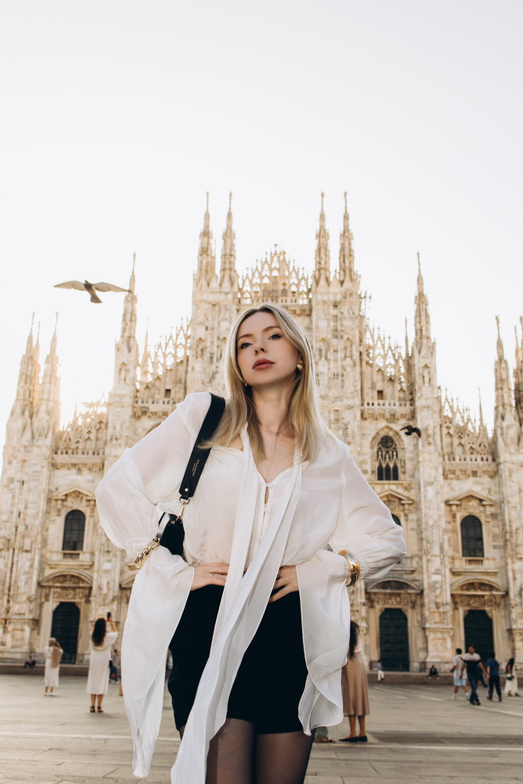 Low-angle portrait of a woman with hands on her hips in a white blouse before the central facade of the Duomo di Milano, a bird flying past the spires