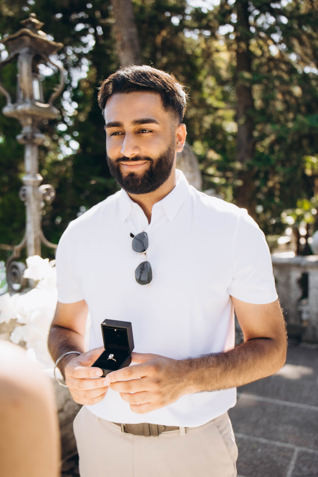 Man in a white polo holding an open ring box with a diamond engagement ring beside an antique lantern on the proposal terrace in Tremezzo