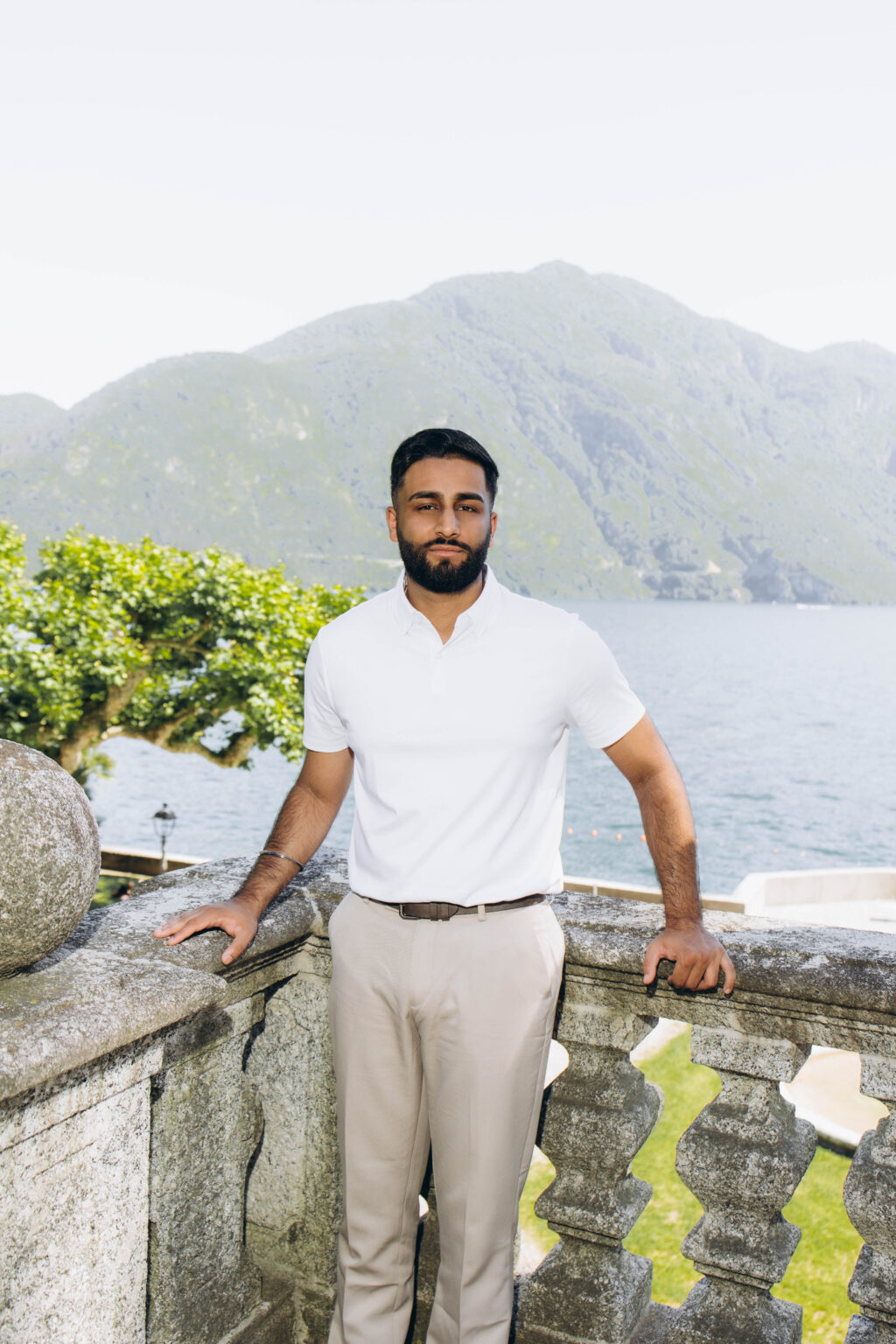 Portrait of the newly engaged man in a white polo leaning on a stone balustrade in Tremezzo, with Lake Como and a mountain behind