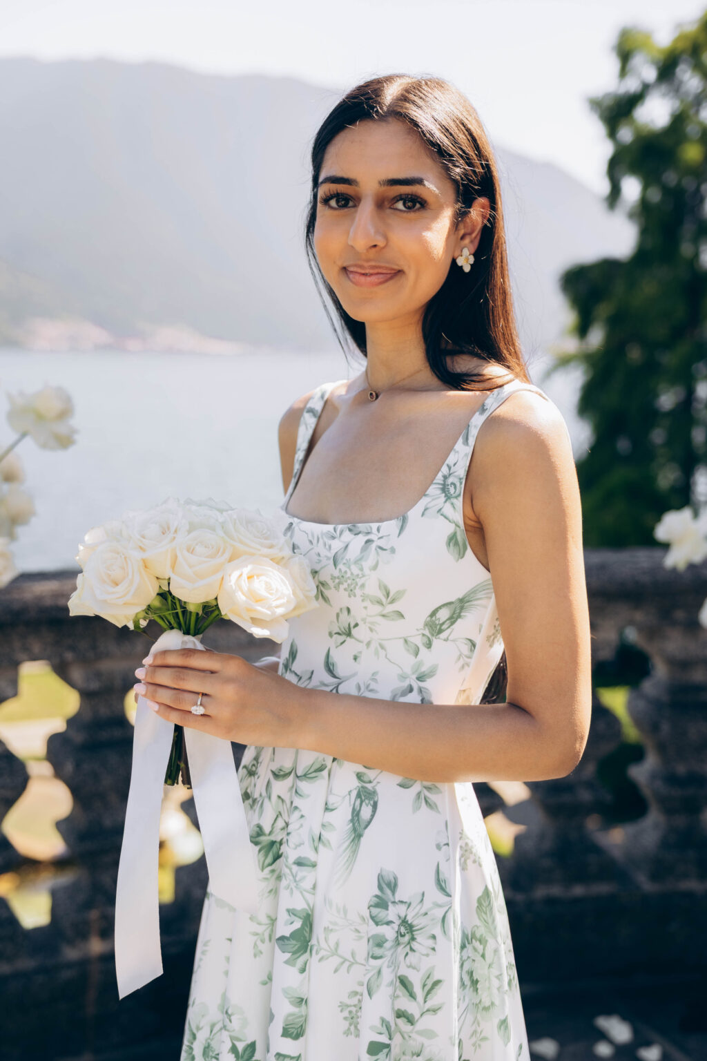 Portrait of the newly engaged woman in a green floral dress holding a white rose bouquet with a satin ribbon, engagement ring visible, Lake Como in the background