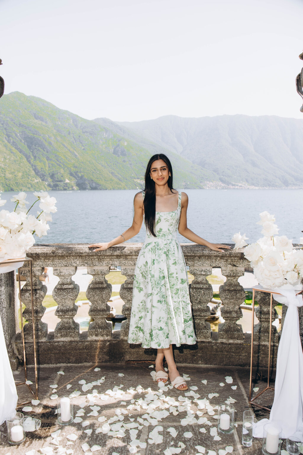 Newly engaged woman standing at the stone balustrade within the white rose proposal setup, with candles, petals, Lake Como, and mountains behind in Tremezzo