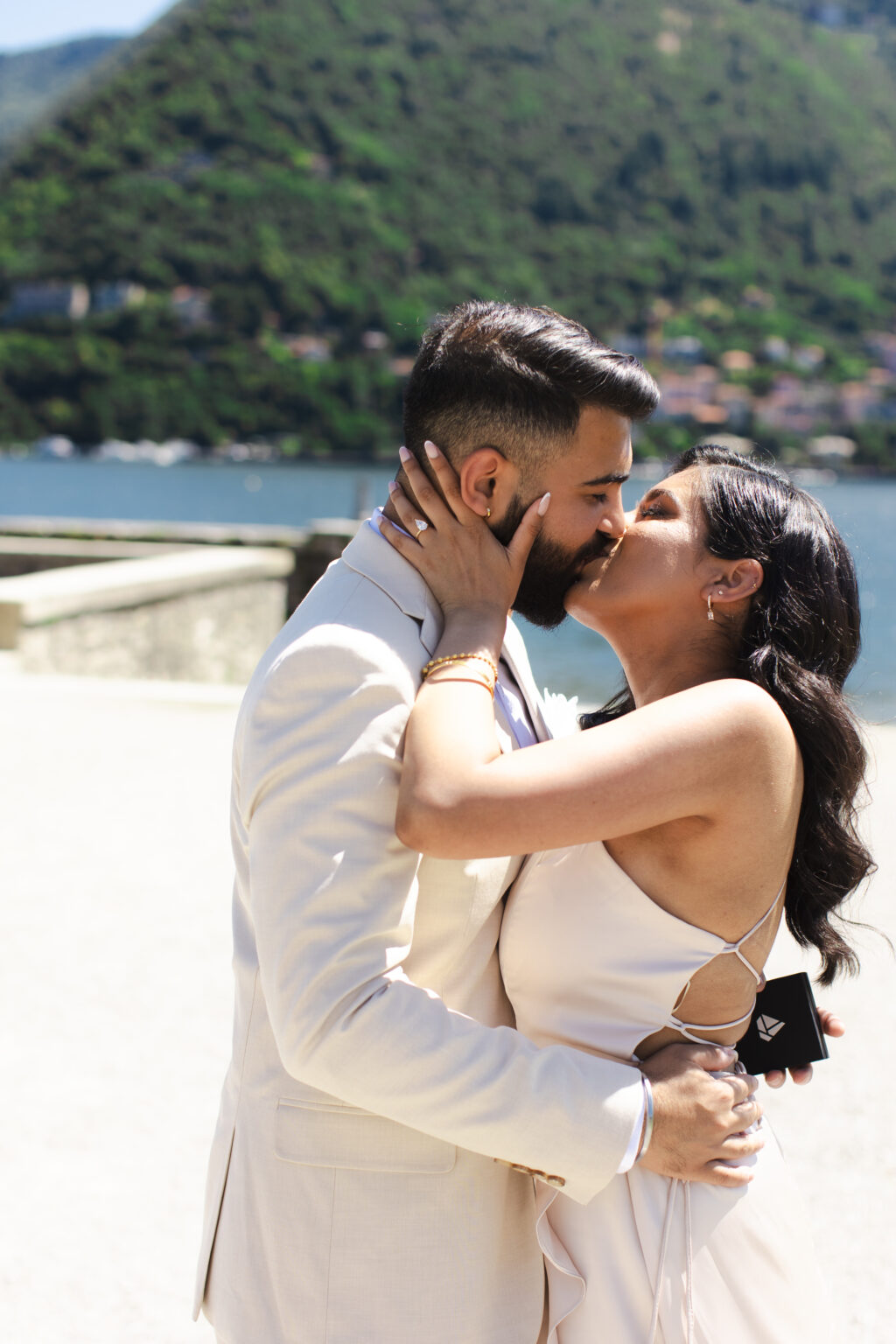 A newly engaged couple sharing their first kiss after saying yes, on a lakeside terrace on Lake Como with the diamond ring on her hand.