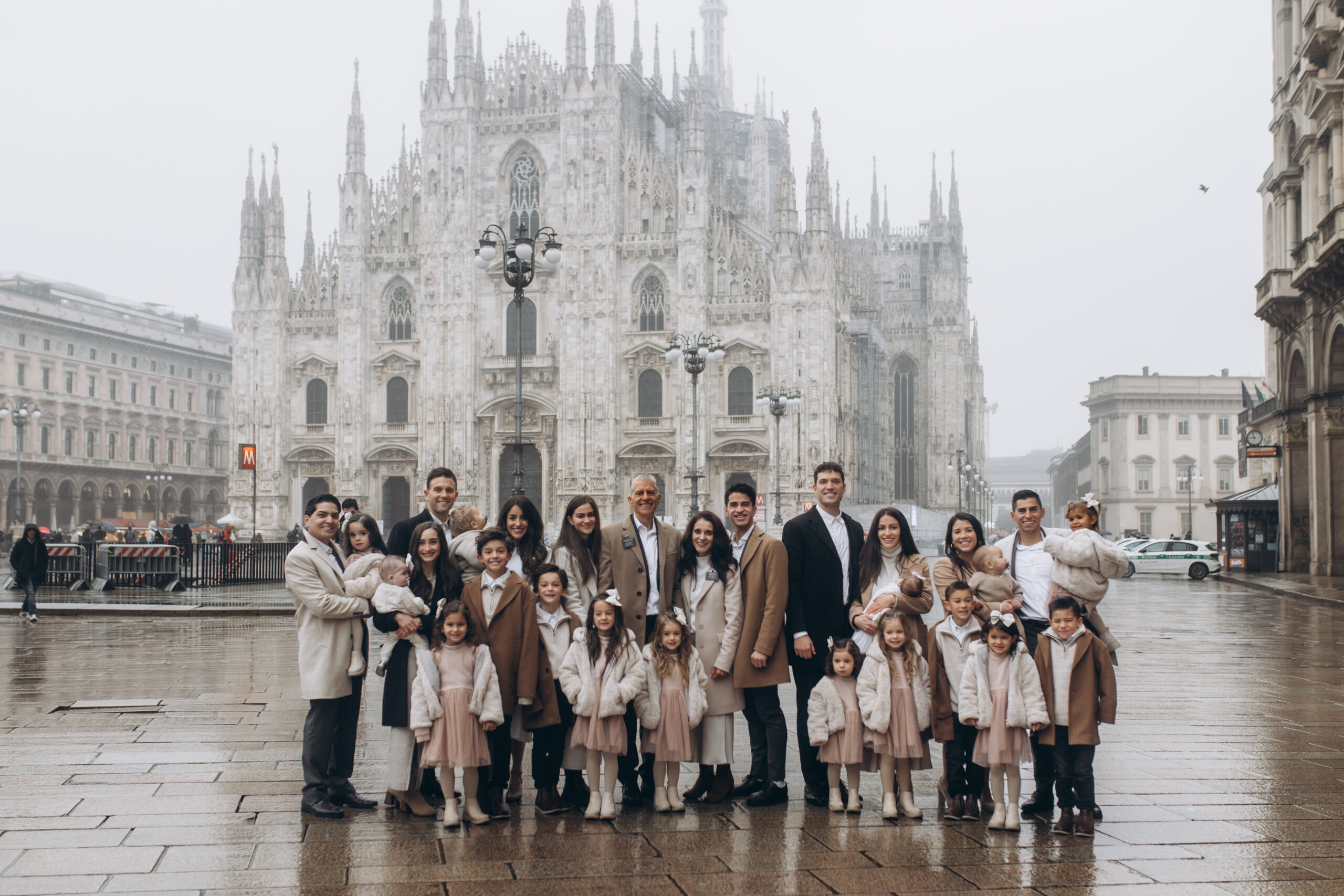 A large group photoshoot in Piazza del Duomo on a misty winter morning, with fog wrapping the cathedral and rain-slicked stone underfoot in Milan. Photographed by Julia, private photographer in Milan. A large family group poses in front of the Duomo cathedral on a foggy, rainy winter day with wet cobblestones in Milan