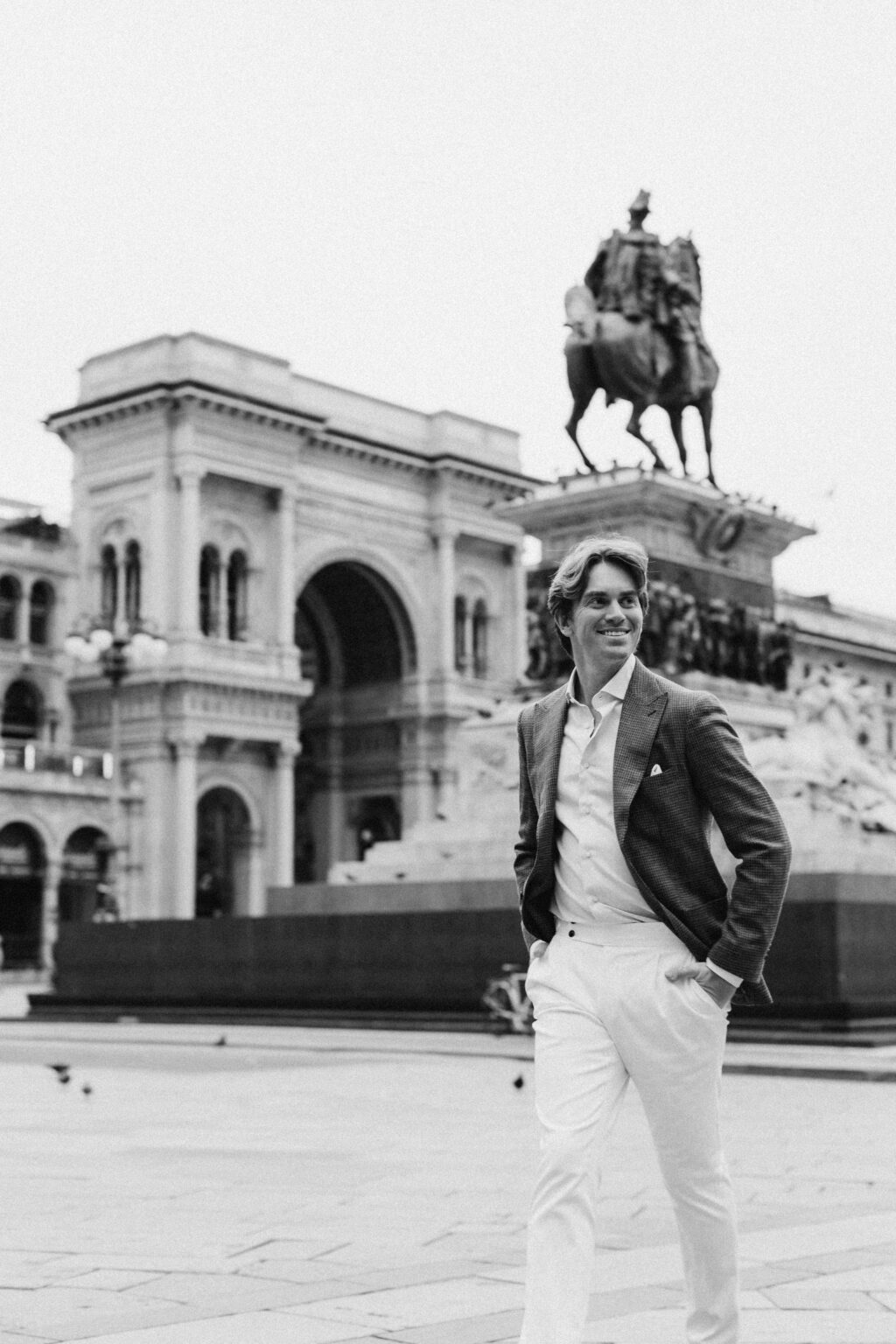 A black and white photo of a smiling man walking in front of the Galleria Vittorio Emanuele II entrance and the Victor Emmanuel monument in Milan.