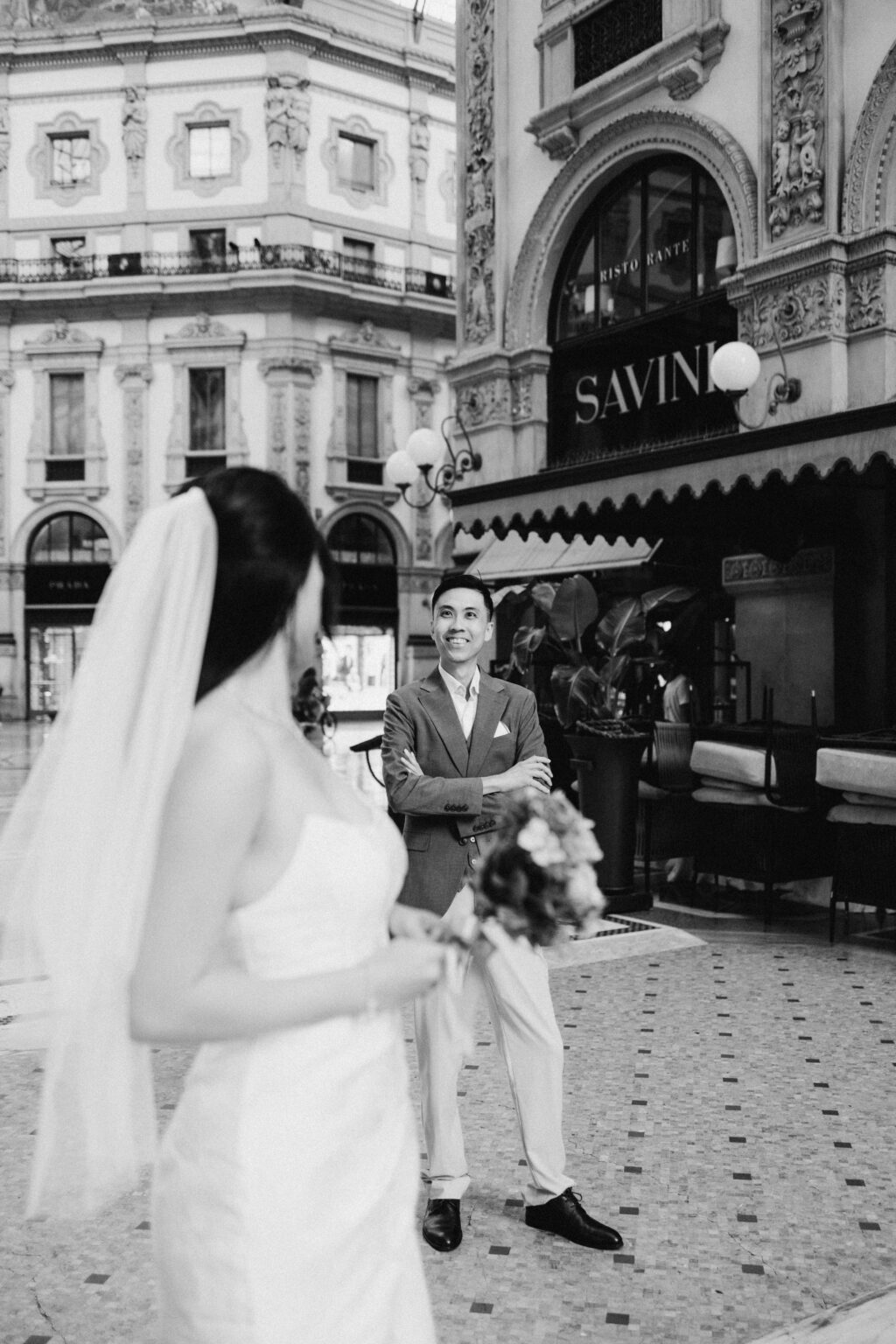 Groom-to-be in a brown jacket standing with arms crossed and smiling at his bride-to-be in the Galleria in Milan, black and white