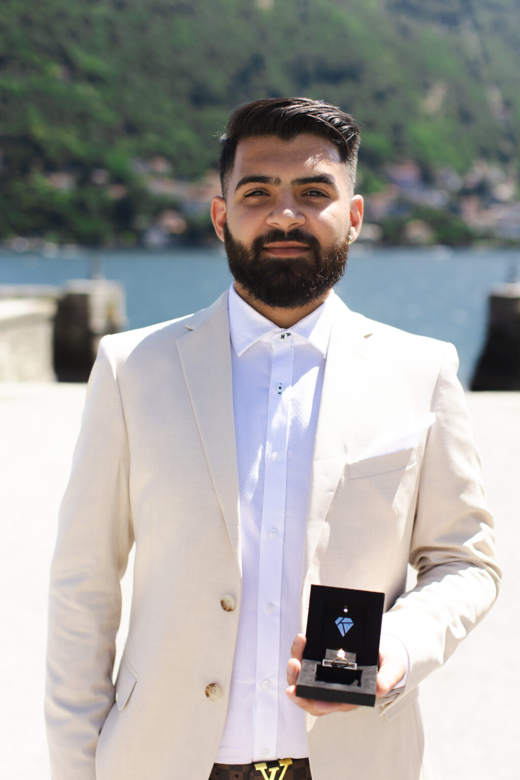 Man in a cream suit holding an open engagement ring box and smiling on a lakefront terrace on Lake Como
