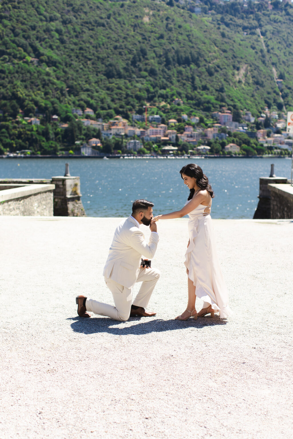 A kneeling man kissing his partner's hand during a proposal on a lakeside terrace on Lake Como, the water and hillside town behind them.