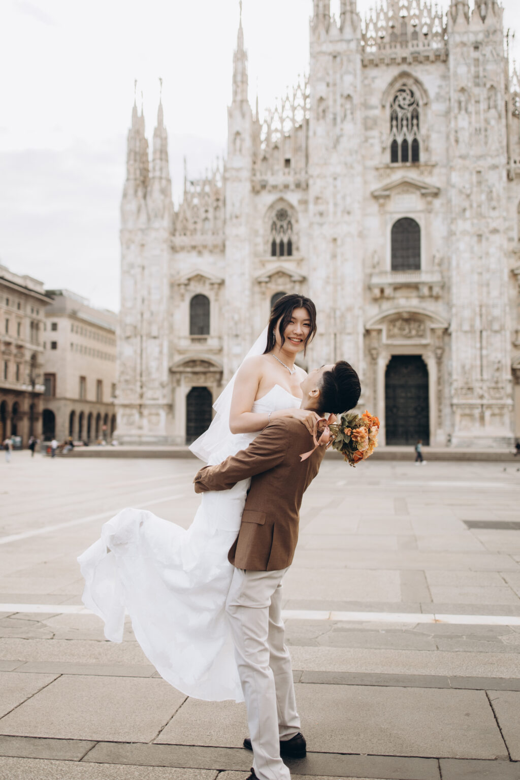 Groom-to-be lifting his laughing bride-to-be into the air with her dress swirling in front of the Duomo in Milan