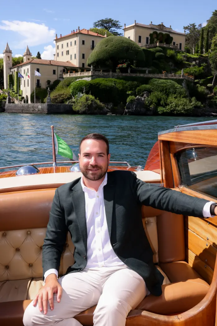 Man in a blazer on a wooden boat with Villa del Balbianello behind him