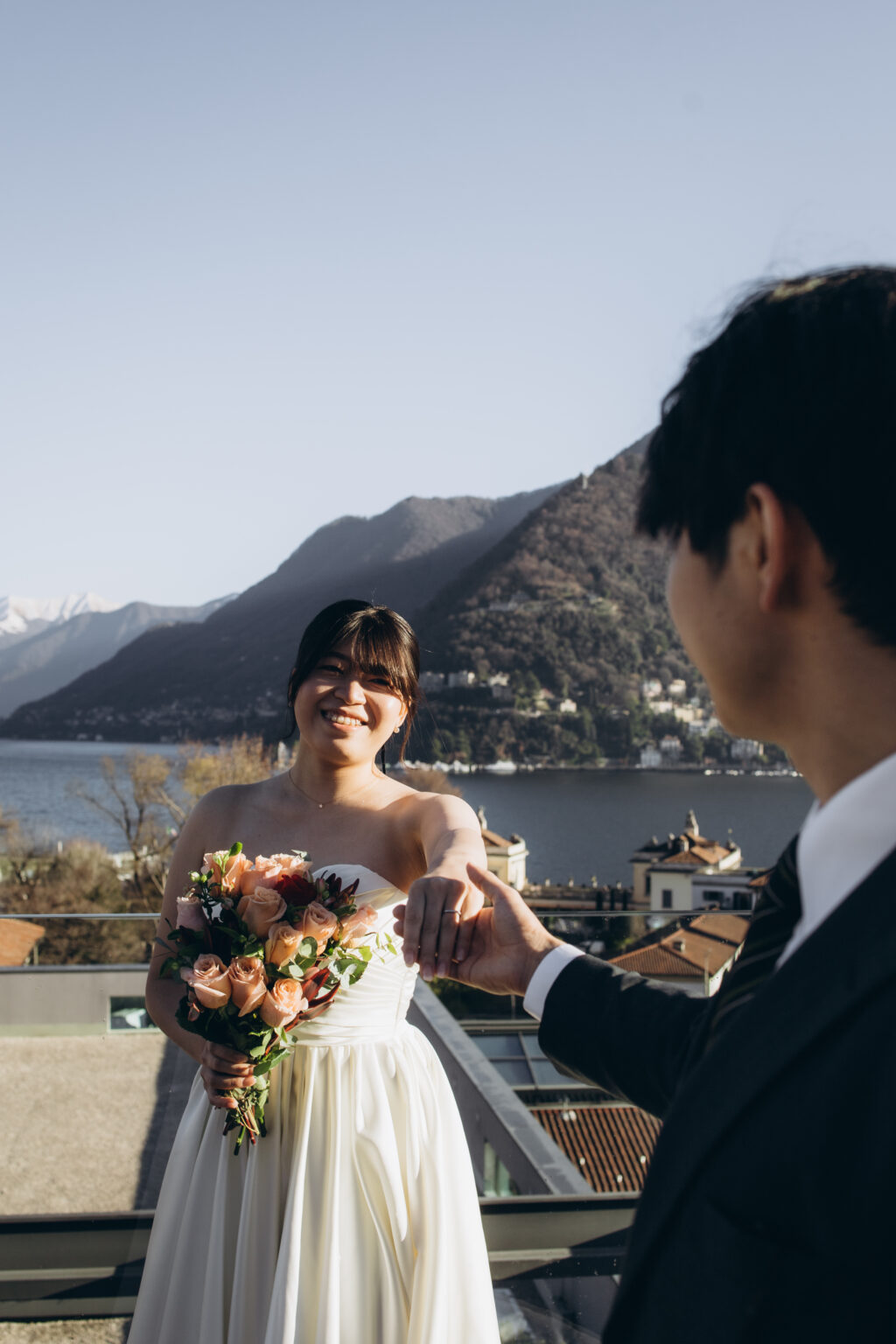 Groom reaching out for his laughing bride's hand on a rooftop terrace with Lake Como and mountains behind her