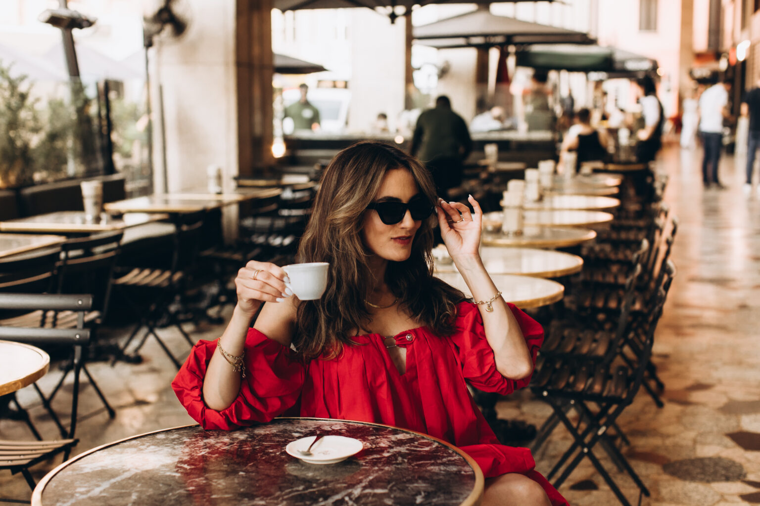 Woman in a red dress and sunglasses drinks espresso at a marble café table in Milan.