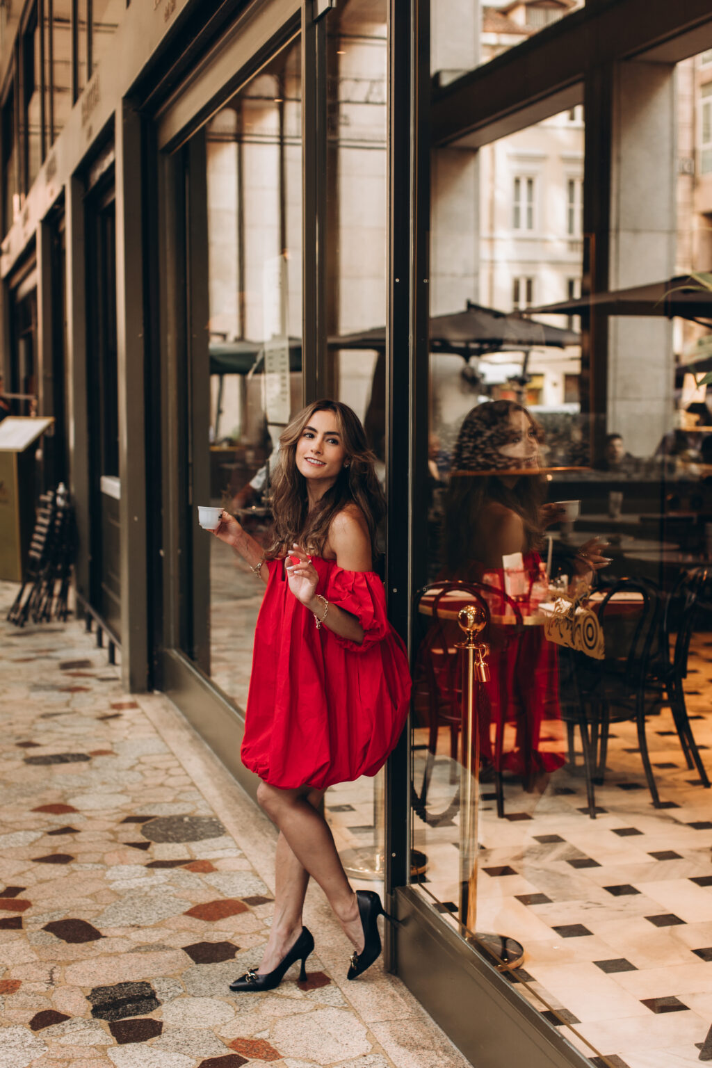 Smiling woman in a red off-shoulder dress leans against a café window with an espresso cup in Milan.