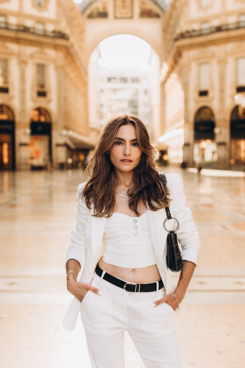 Woman in a white blazer and crop top poses before the blurred arch of the Galleria Vittorio Emanuele II in Milan.