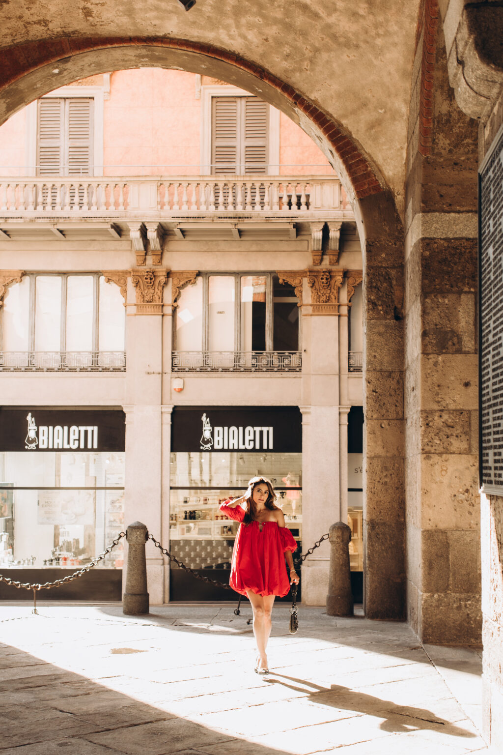 Woman in a red dress walks through a stone arch of the Palazzo della Ragione arcade on Piazza dei Mercanti in Milan.