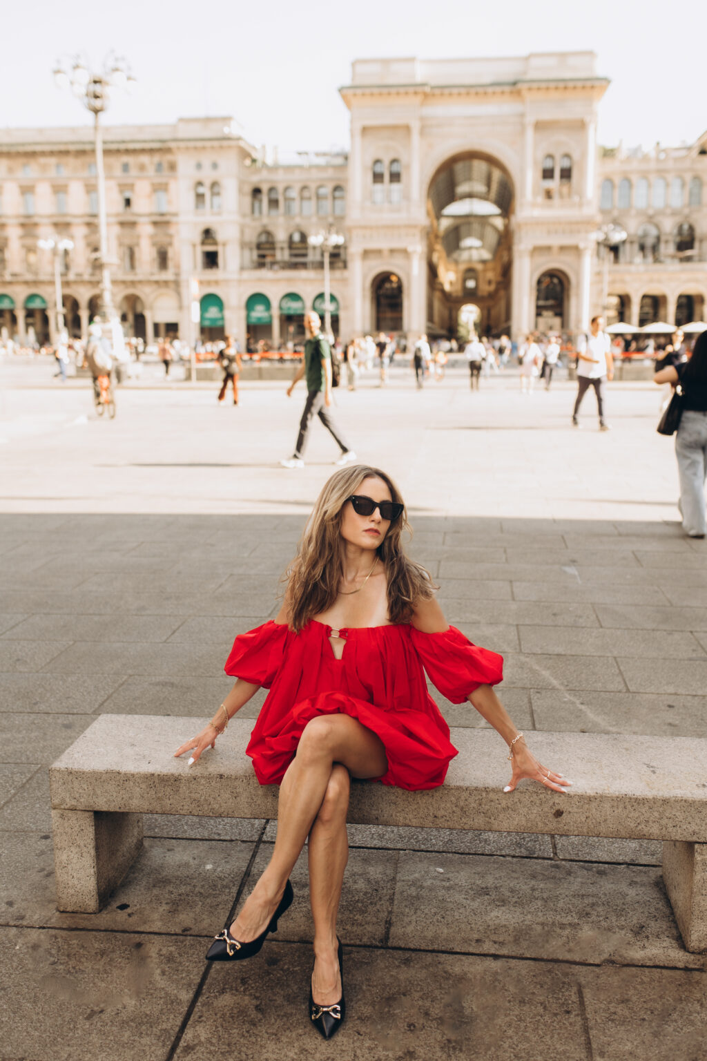 Woman in a red dress sits on a stone bench in Piazza del Duomo with the triumphal arch of the Galleria Vittorio Emanuele II behind her in Milan.