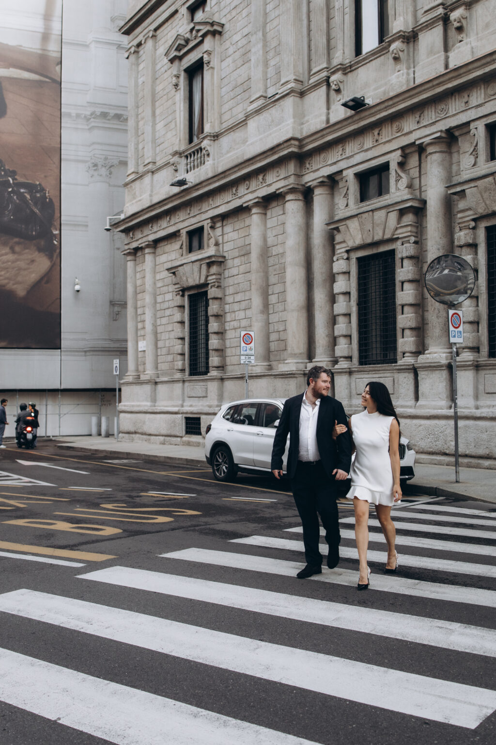 Just-engaged couple in a black suit and white dress walking arm in arm across a pedestrian crossing on a historic street in central Milan