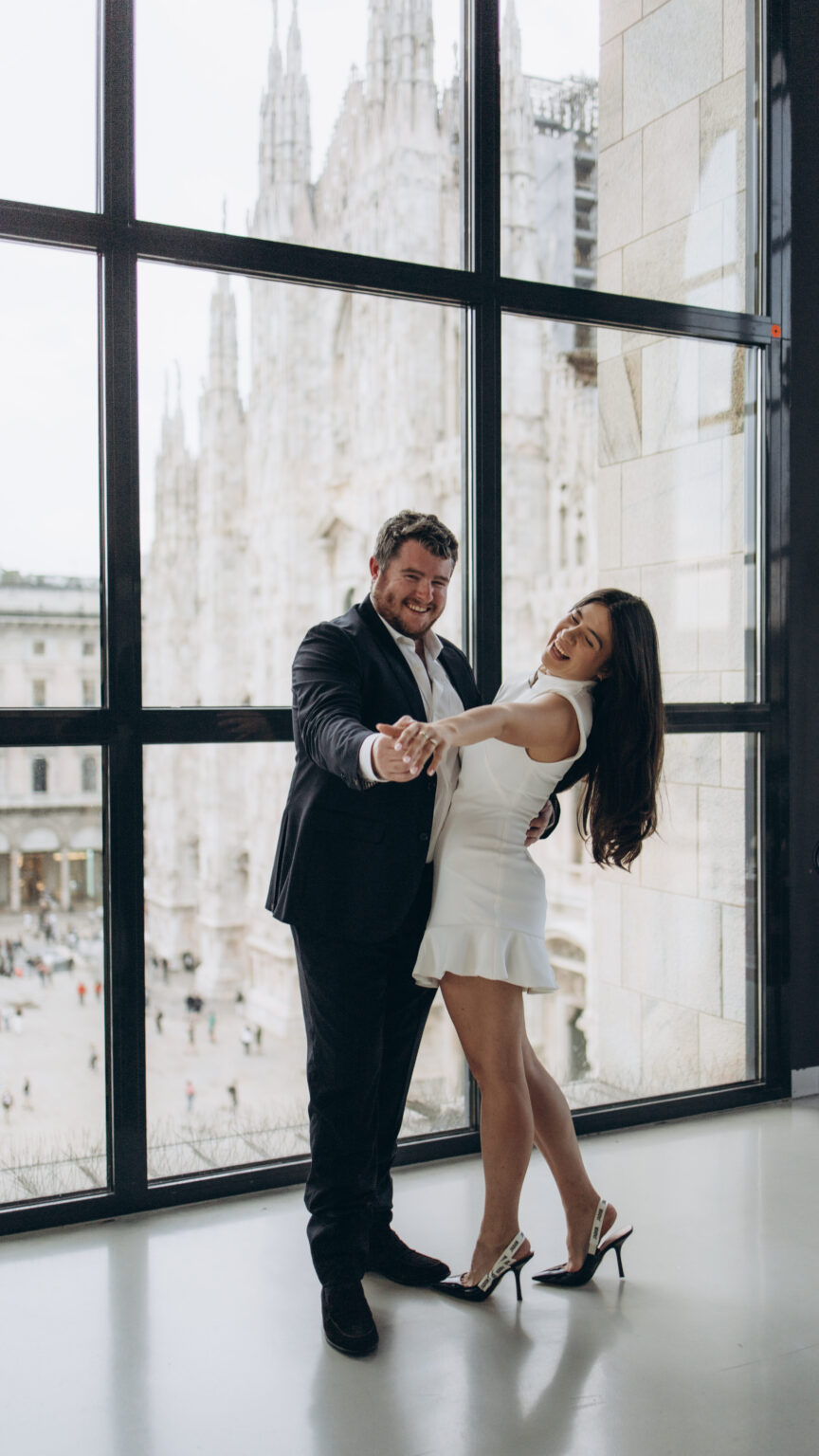 Just-engaged couple laughing and dancing playfully in front of the Museo del Novecento window overlooking the Duomo in Milan
