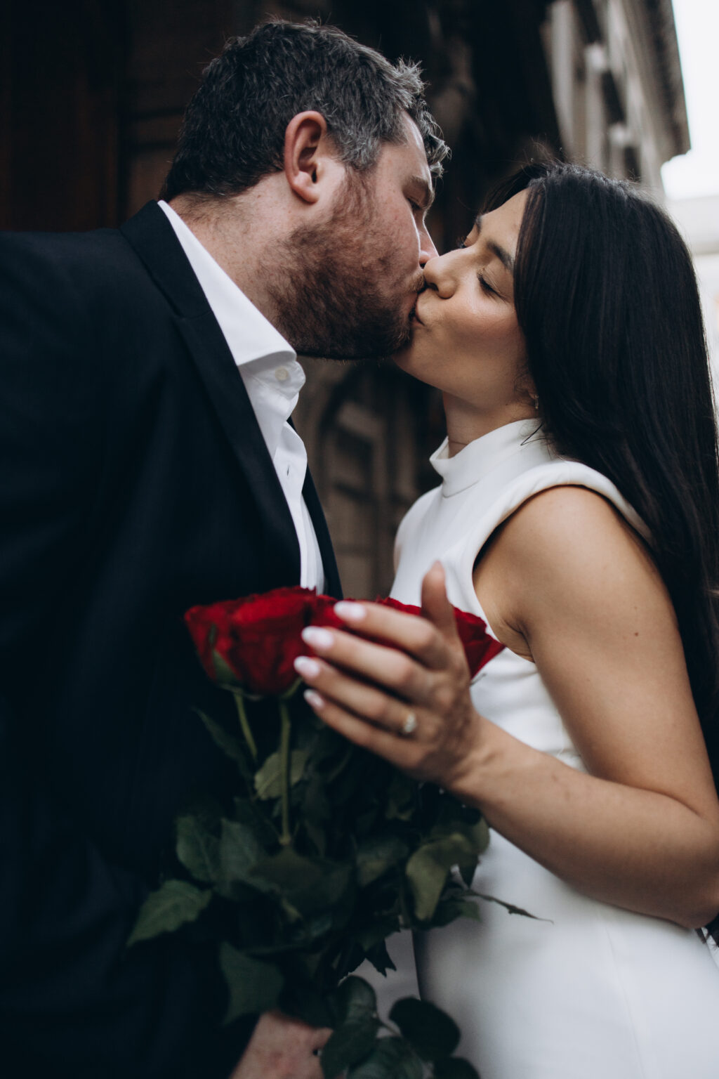 Newly engaged couple kissing while holding a bouquet of red roses in the dark ornate doorway of the Casa degli Omenoni in Milan