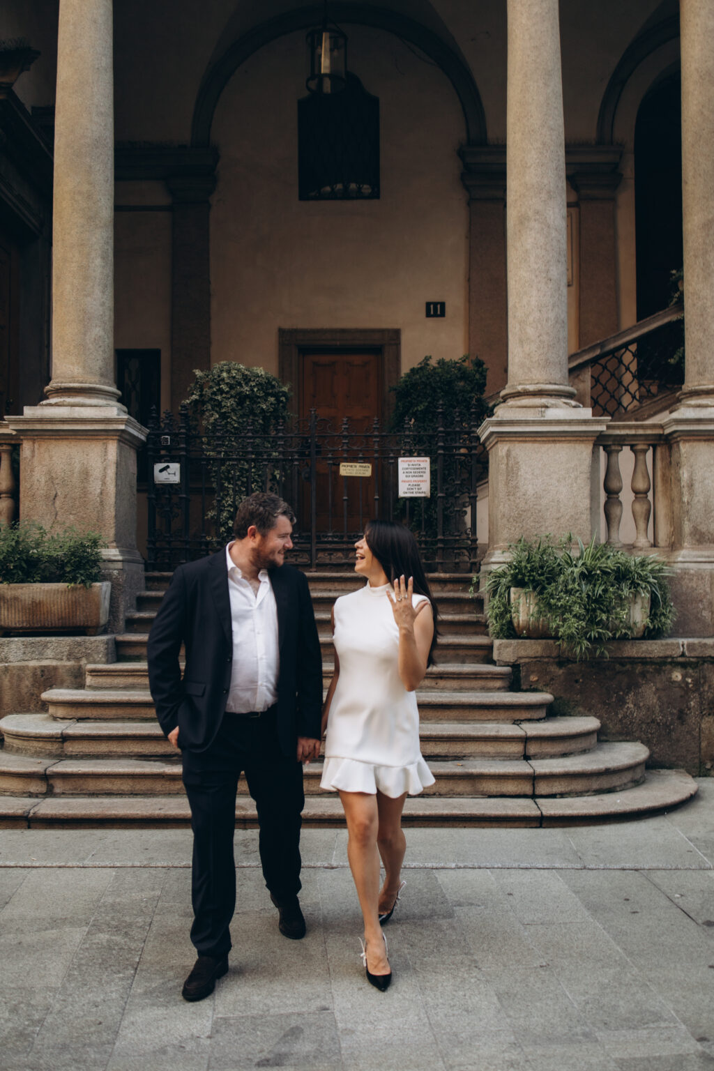 Just-engaged couple walking hand in hand and laughing together in front of a columned staircase in a historic Milan courtyard