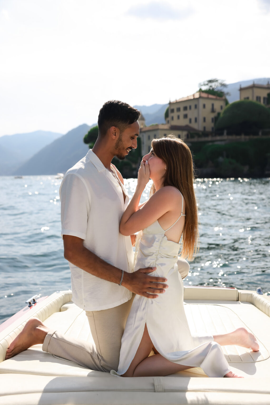 A newly engaged couple leans close in a tender moment on a boat, with the Villa del Balbianello promontory behind them on Lake Como.