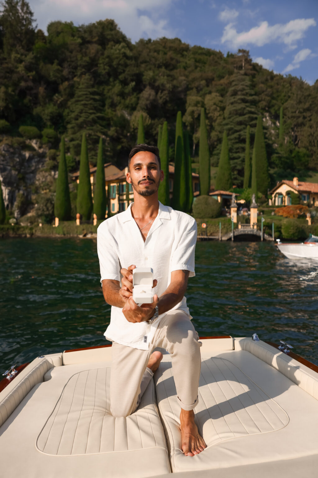 A man kneels on the bow of a boat and holds open a ring box toward the camera, with an ochre lakeside villa and tall cypresses behind him on Lake Como.
