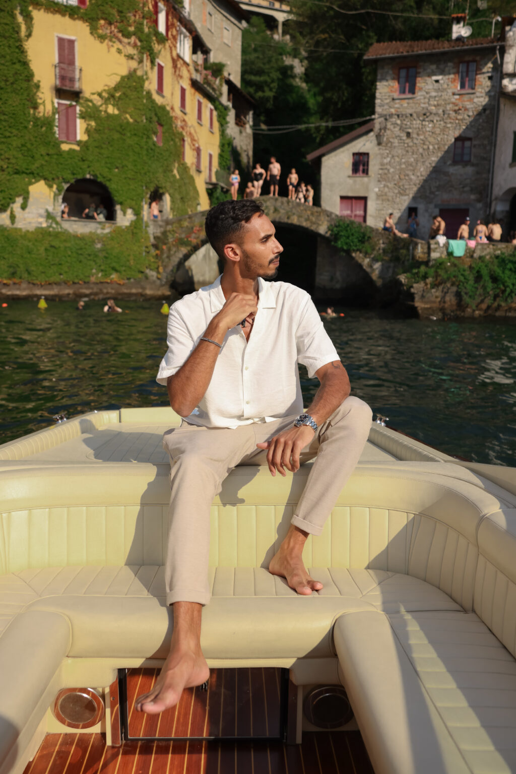 A man in a white linen shirt sits alone on a boat before the medieval Ponte della Civera bridge in Nesso, on Lake Como, at the start of a proposal photoshoot.