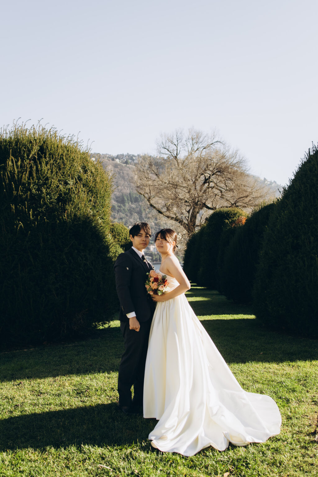 Bride in a white gown with a bouquet standing beside her groom in a topiary-lined garden allee overlooking Lake Como and the Brunate hillside