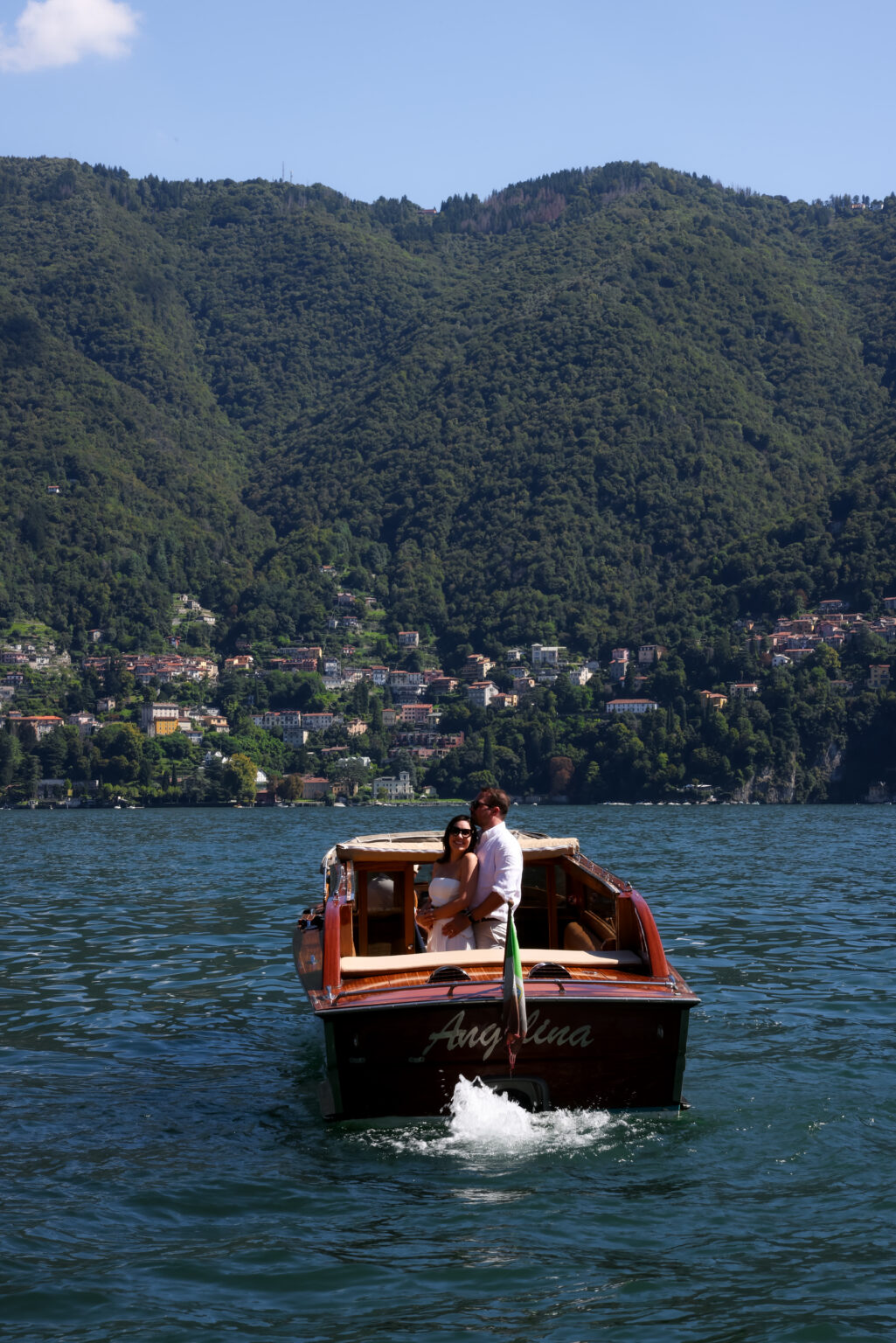 Couple embracing at the stern of a classic wooden boat named Angelina on Lake Como, with the green hills and lakeside town of Como behind them