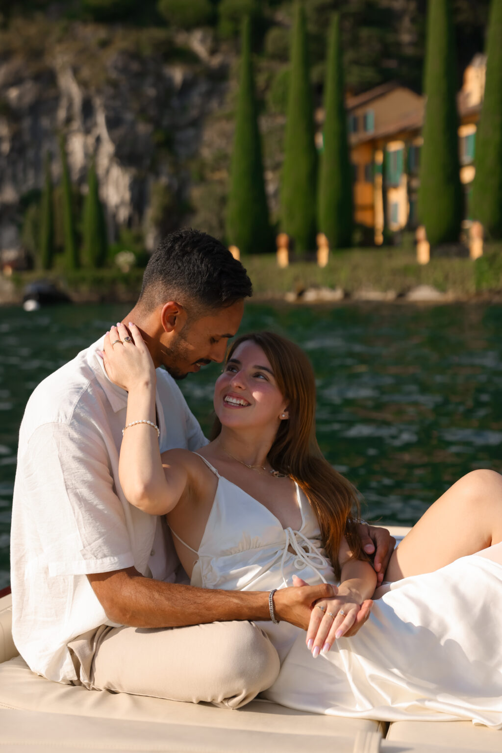 A newly engaged woman smiles up at her fiancé as they embrace on a boat before an ochre villa and cypresses on Lake Como.