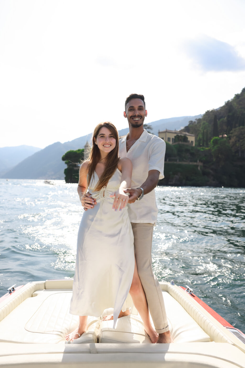 A newly engaged couple stands on the bow of a boat smiling at the camera as the woman shows her ring, with the wooded western shore of Lake Como behind them.