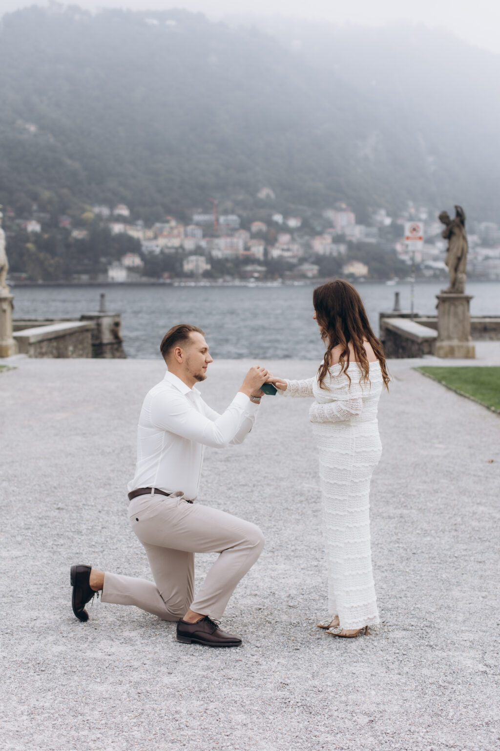 He kneels and holds her hands as she accepts the ring, a neoclassical angel statue and the misty lake behind them, in the gardens of Villa Olmo on Lake Como.