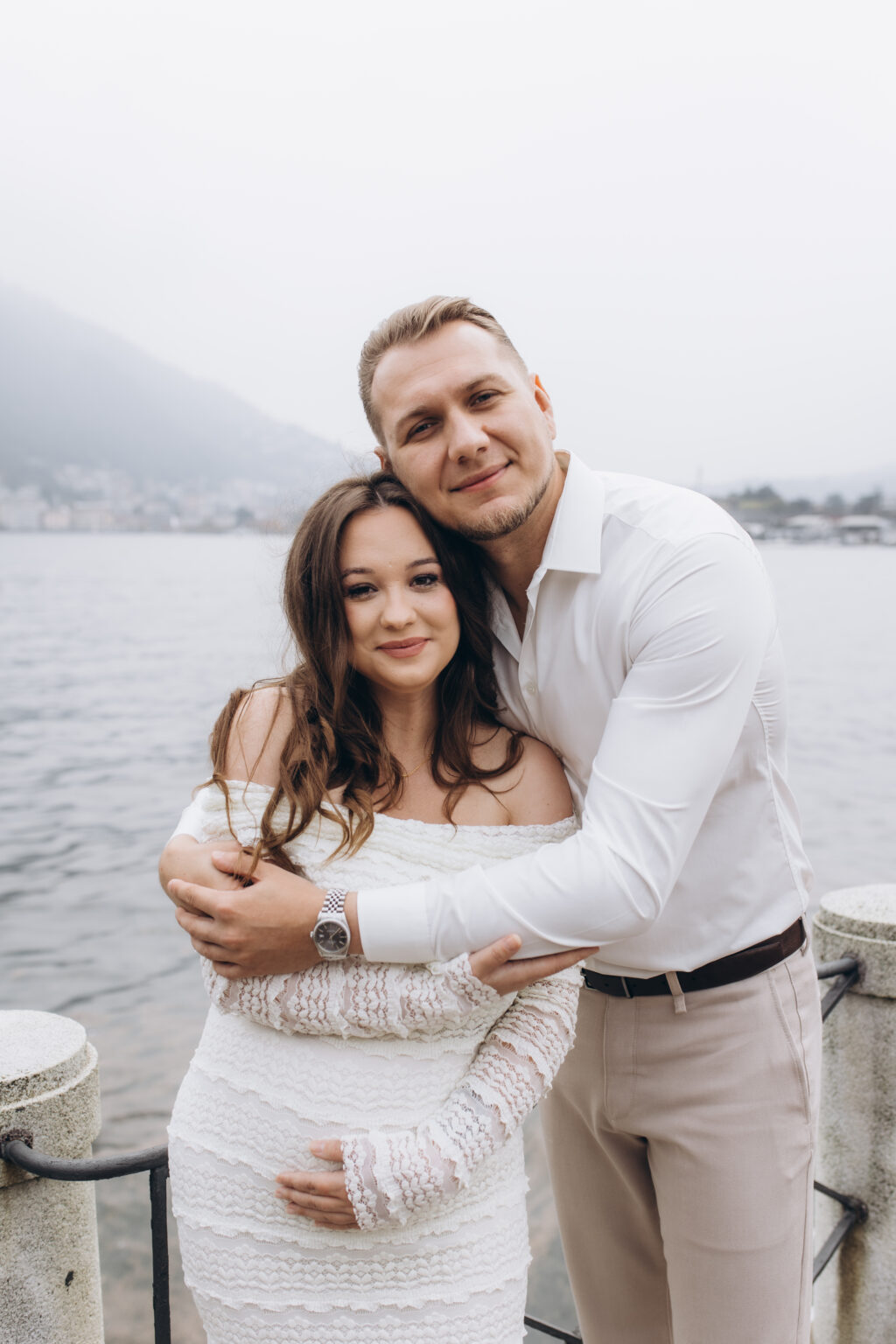 her leans back into his embrace at the Como lakefront railing, both smiling at the camera, the misty lake and town behind them, on Lake Como.