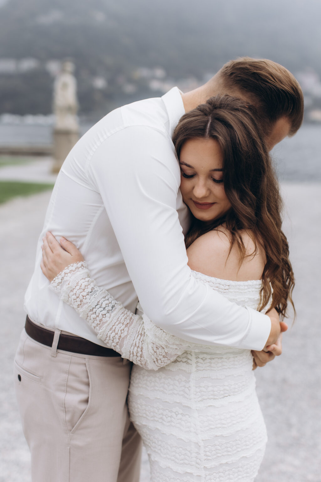 her rests her face against his chest in a close embrace, the misty gardens of Villa Olmo behind them, on Lake Como.